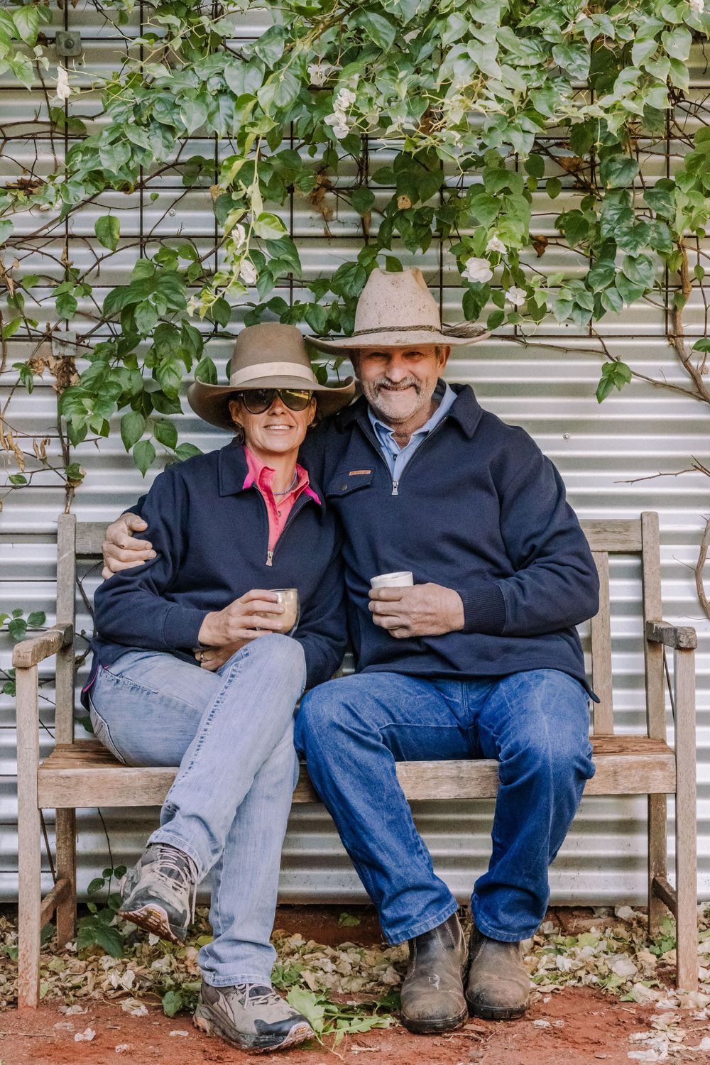Couple in matching navy jackets and cowboy hats share drinks while sitting on garden bench.