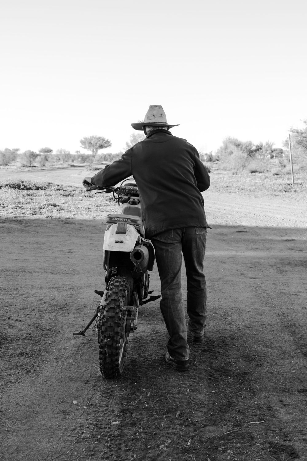 Person in cowboy hat stands beside dirt bike motorcycle on dusty trail.