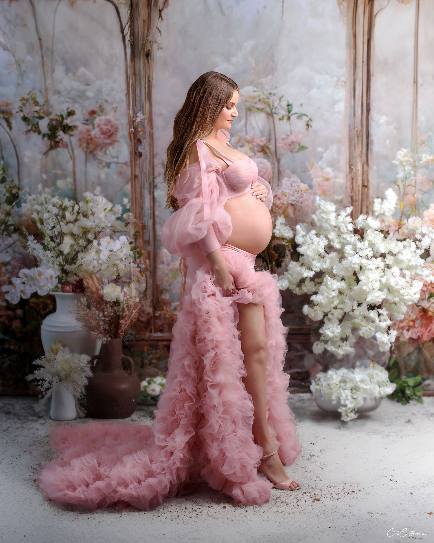 maternity photo of woman wearing peach coloured couture blouse and ruffle skirt against glasshouse backdrop