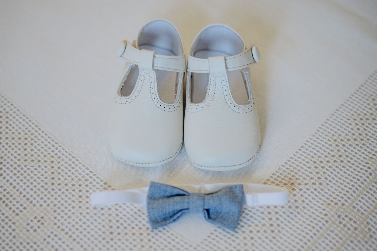 White baby T-strap shoes with light blue bowtie accessory on lace background.