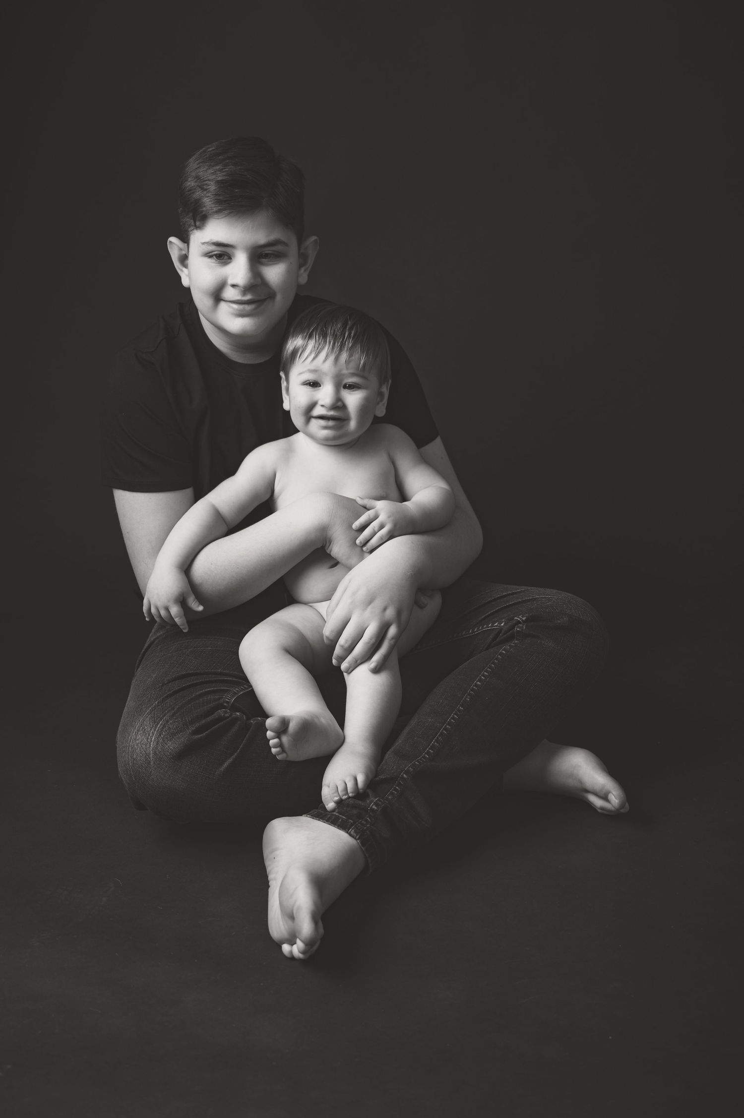 Black and white studio portrait of two siblings sitting together against dark background.