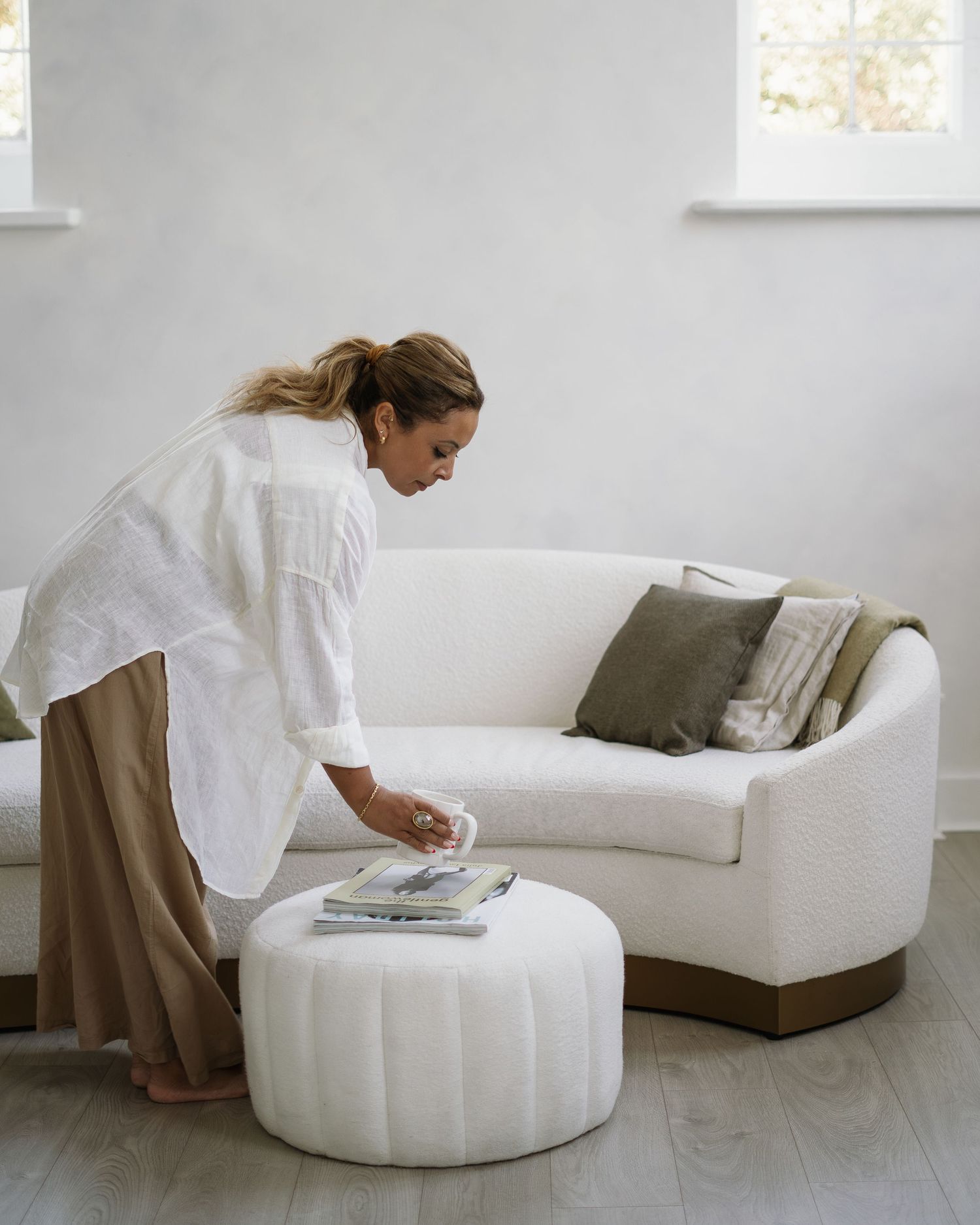 Person in white shirt and brown skirt setting items on a round white ottoman beside a cream colored sofa.