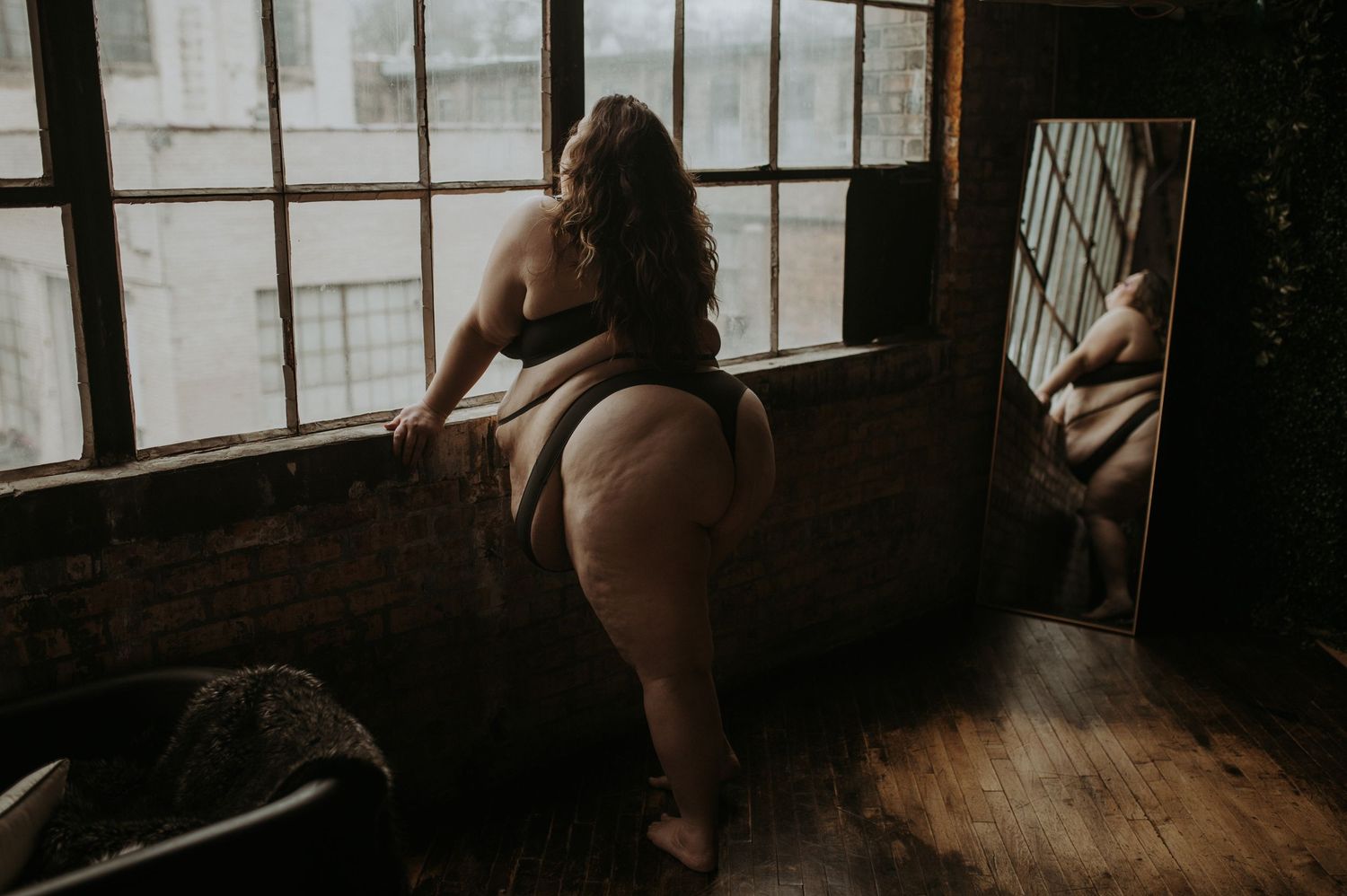 Person wearing black lingerie poses near window in moody boudoir photoshoot with brick wall background.