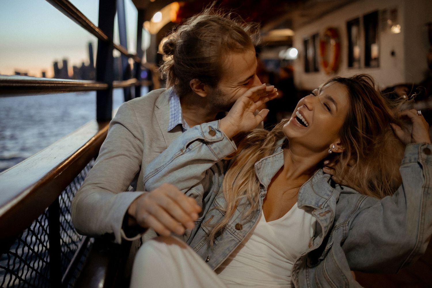 Couple sharing joyful laughter together on boat deck during golden hour with city skyline in background.