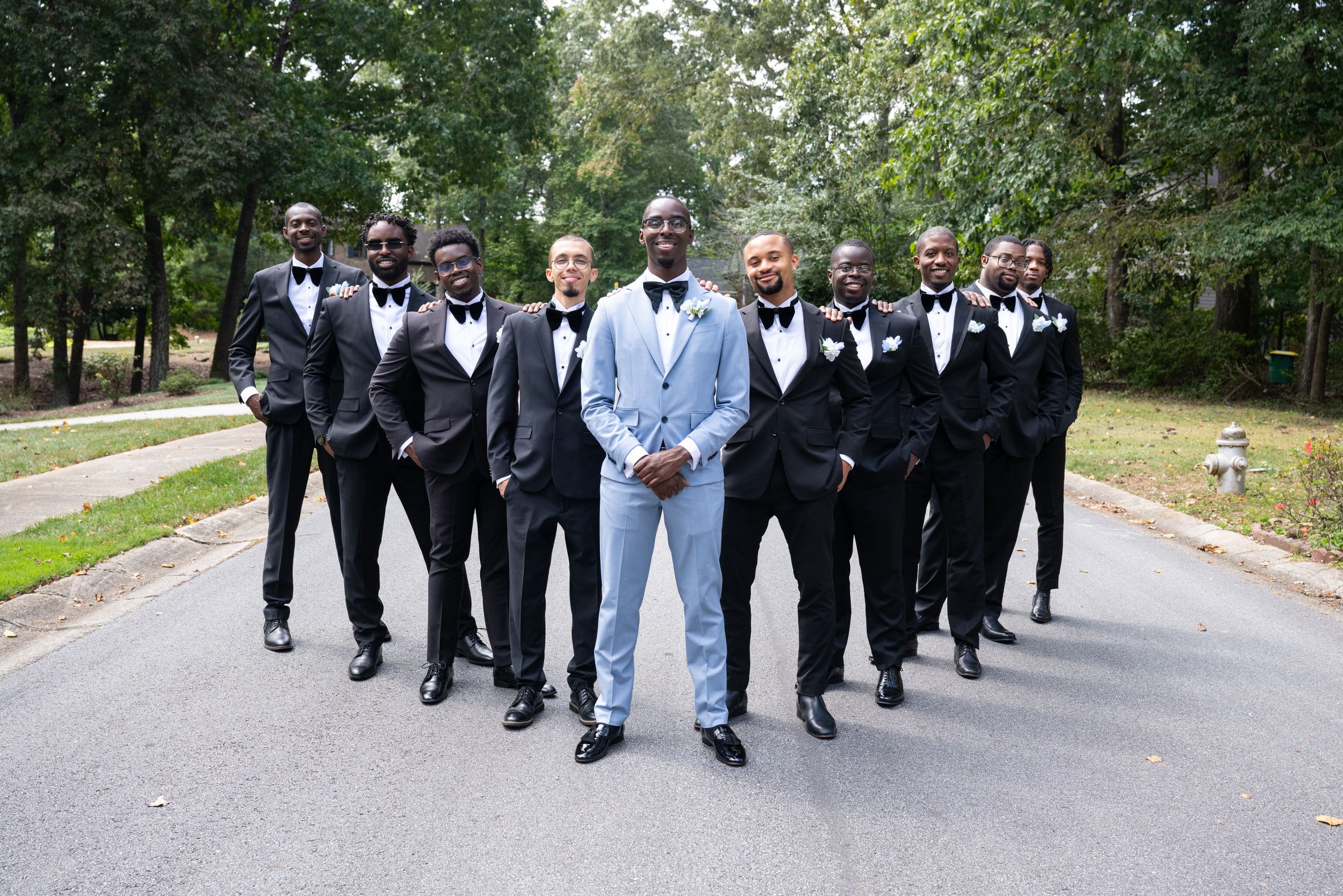 Wedding party in formal attire poses stylishly on tree-lined path.