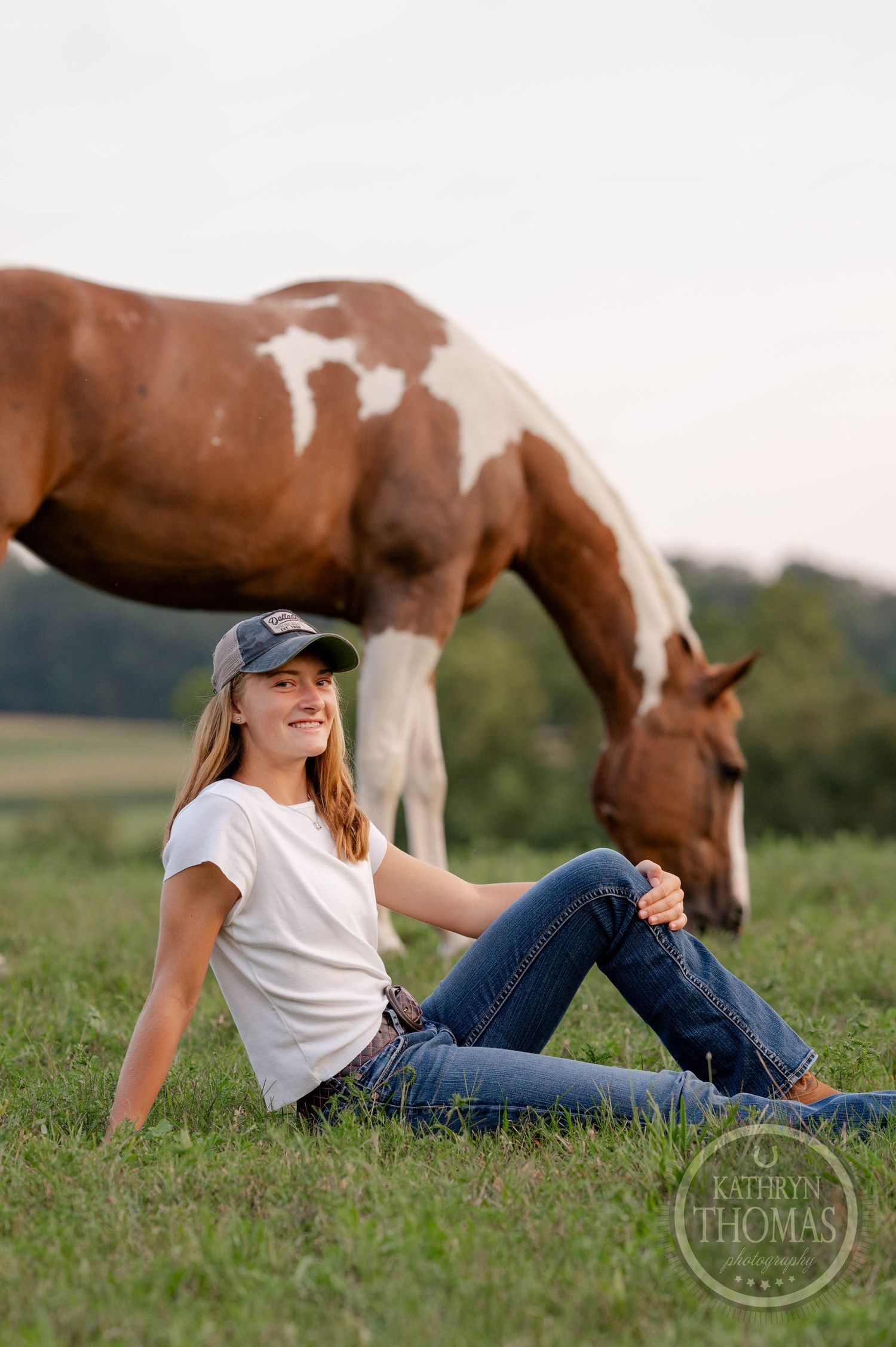 Paint horse bends down to graze while companion in jeans and t-shirt sits on grassy hill.