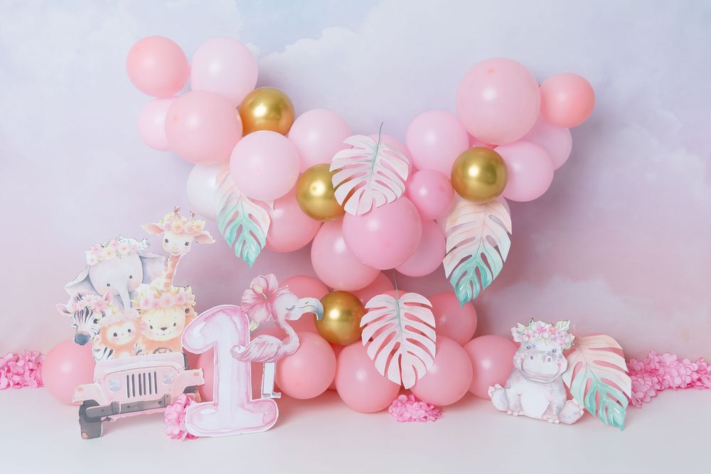 Pink and gold balloon arch with tropical leaves and birthday party decorations in soft pastel colors against light background.