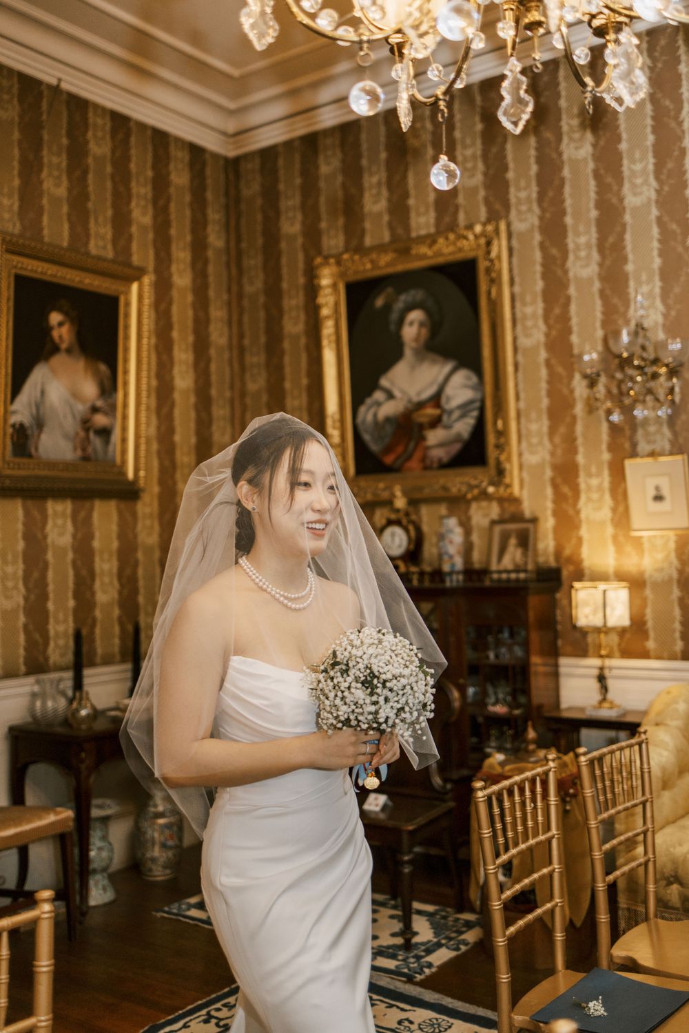 A bride in white dress holds bouquet in elegant room with chandelier and vintage portraits.