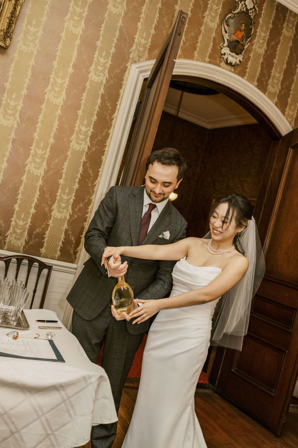 Newlyweds pour champagne together in an elegant vintage room with striped wallpaper.
