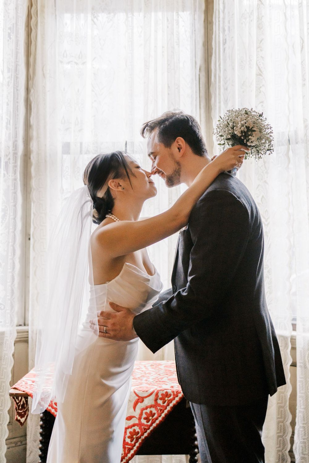 A romantic moment between newlyweds by sunlit windows in an elegant room.