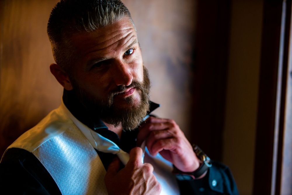 Bearded figure in formal attire gazes thoughtfully by window light in moody indoor setting.