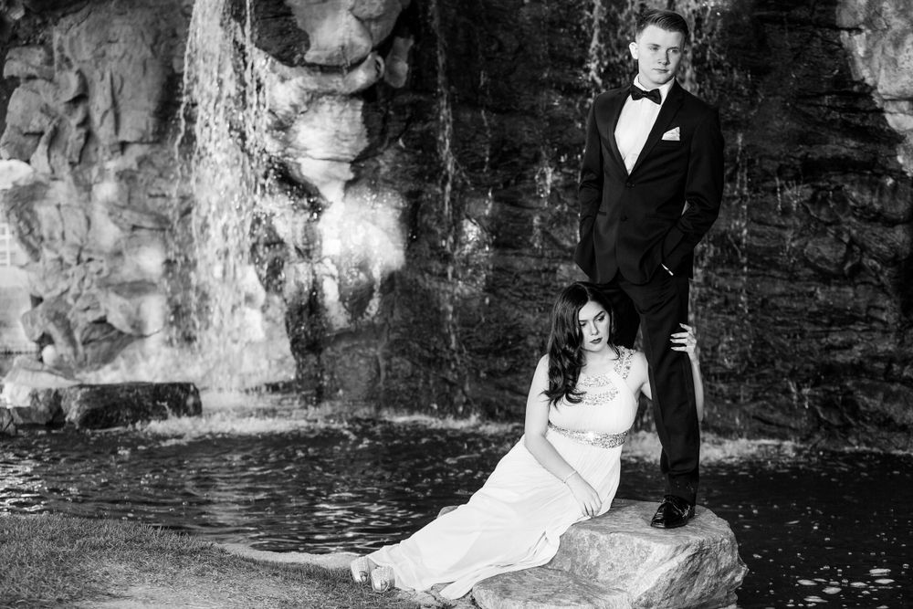 A bride in a white dress sits on rocks near a waterfall while her groom stands beside her in black and white photography.