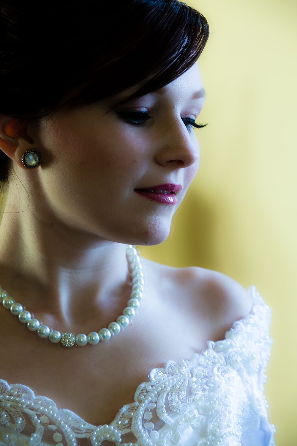 A close-up profile shot showing an elegant pearl necklace and white lace bridal gown against a soft yellow backdrop.