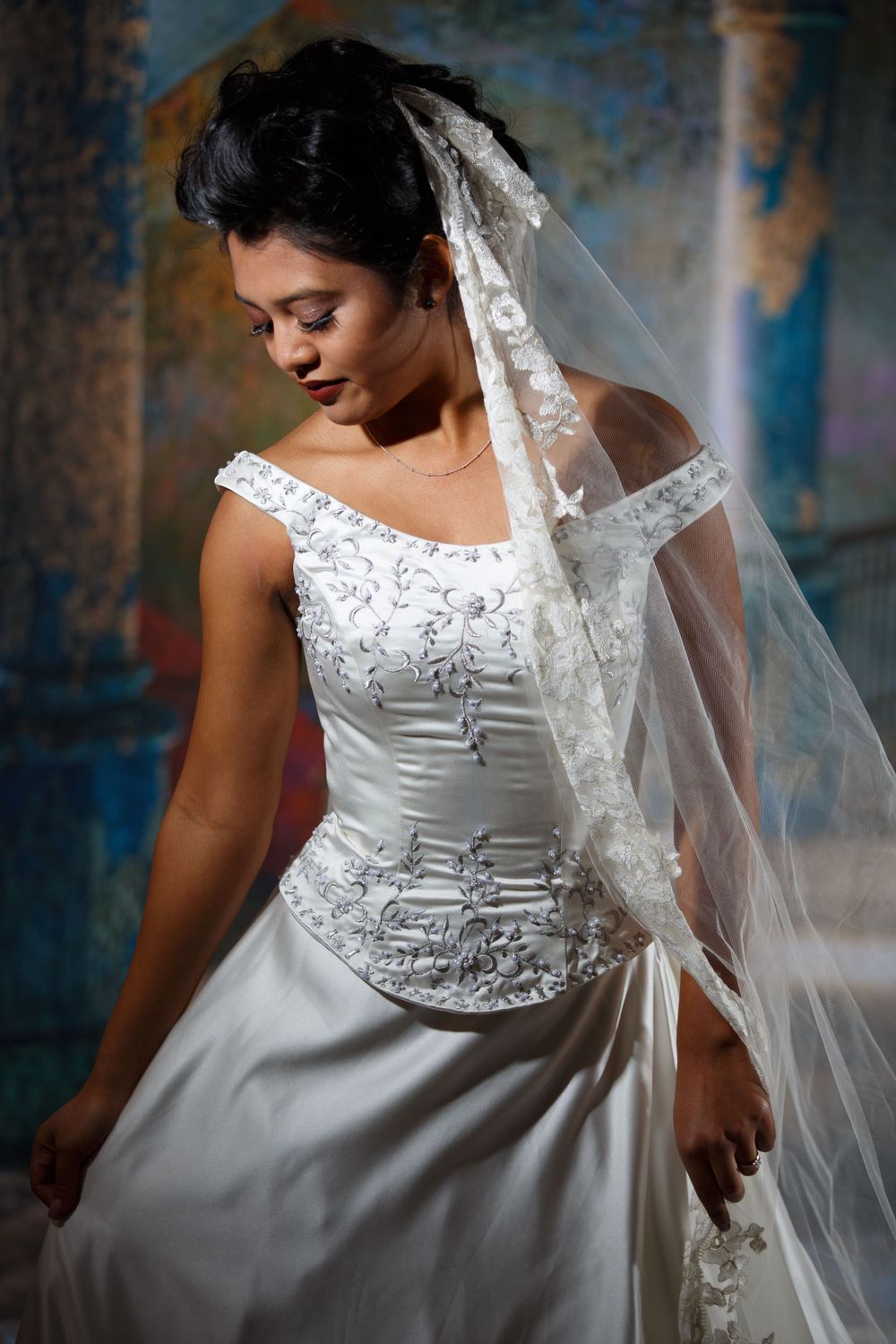 Elegant wedding gown with detailed beaded bodice, off-shoulder neckline and flowing veil against blue textured backdrop.