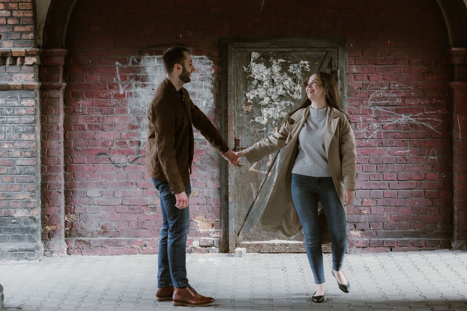 Two people stand holding hands under a brick archway in a romantic urban setting.