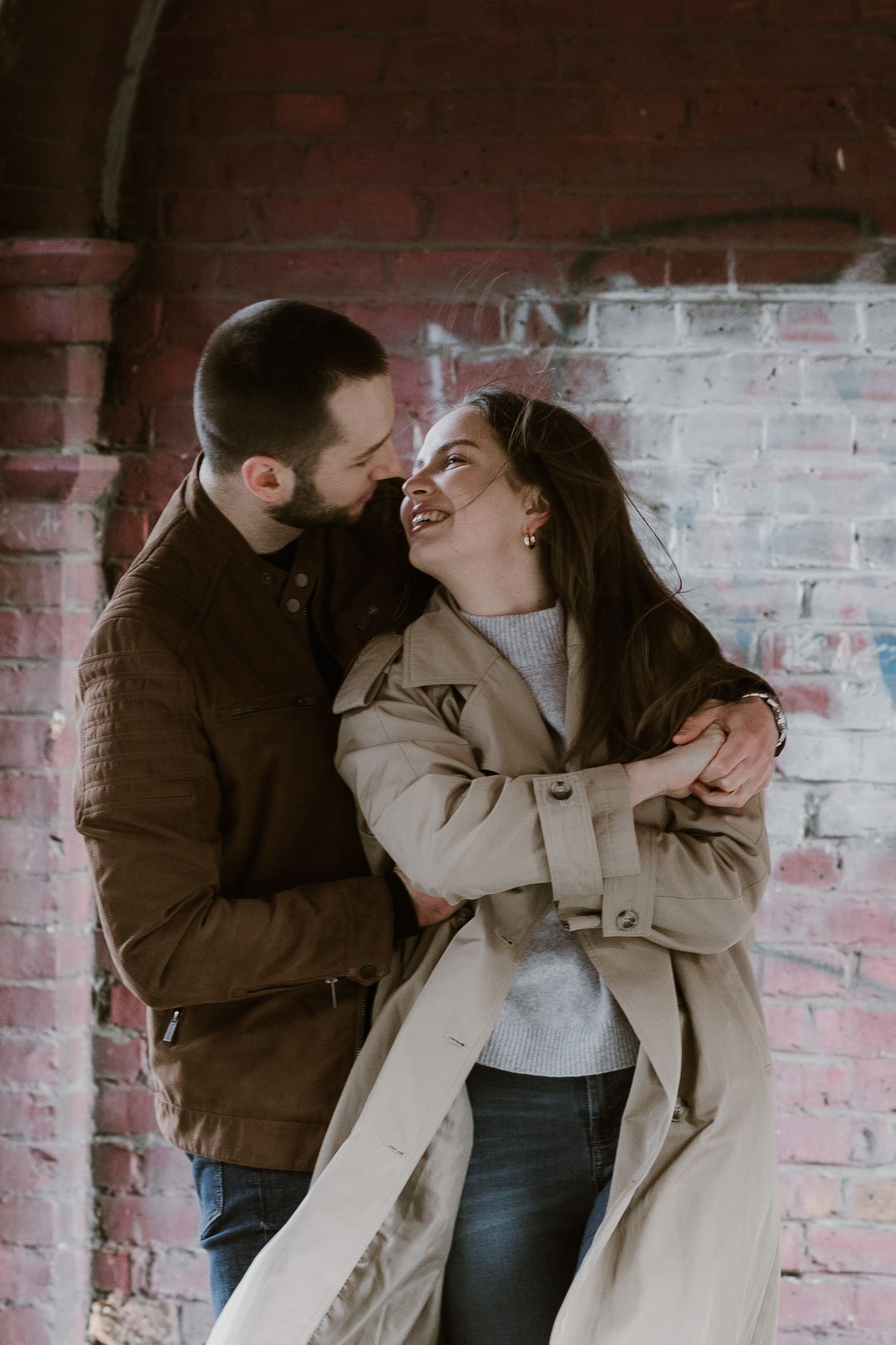 A romantic couple shares a tender moment while leaning against a rustic pink brick wall.