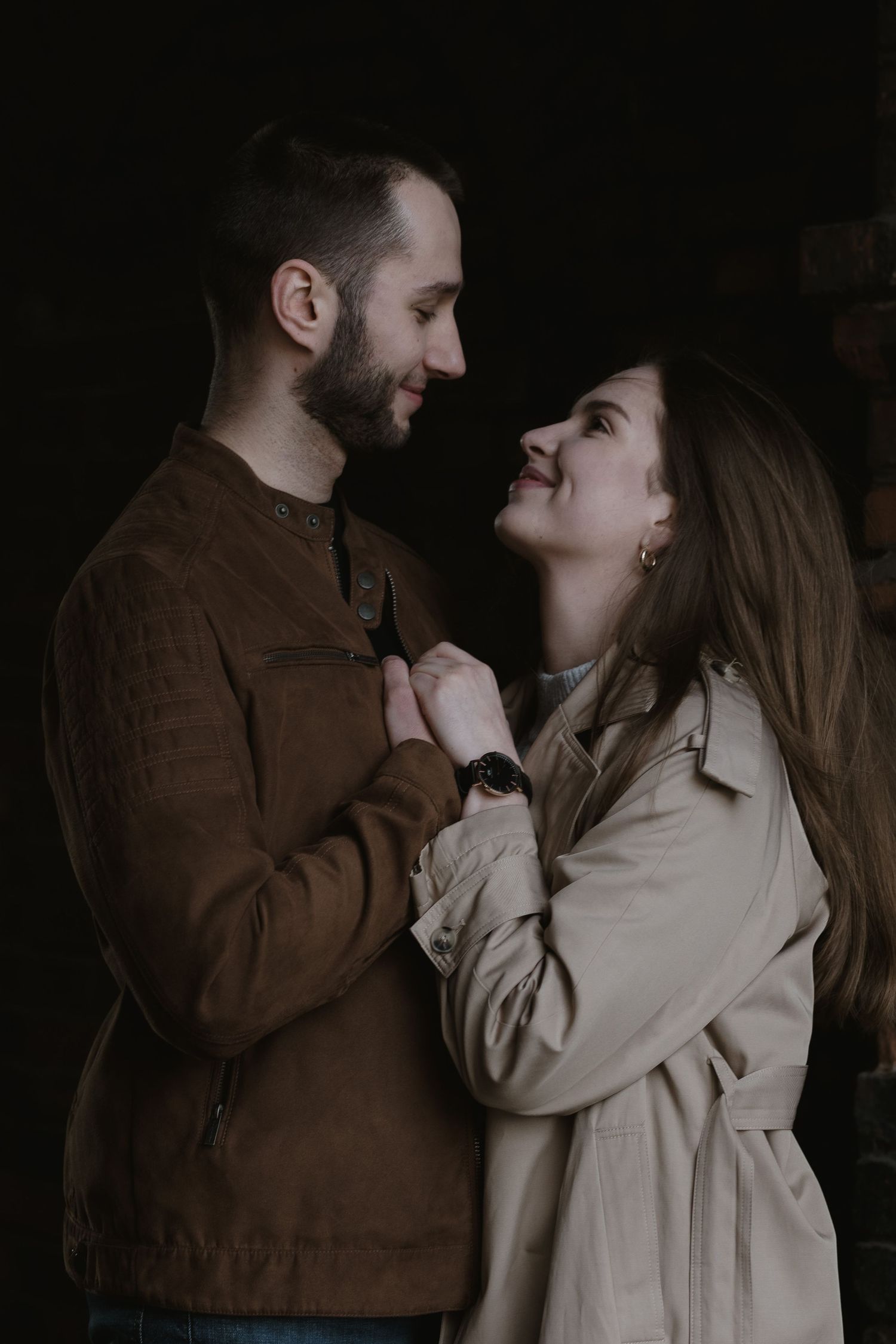A couple shares an intimate moment together in dim lighting wearing a brown jacket and beige coat.