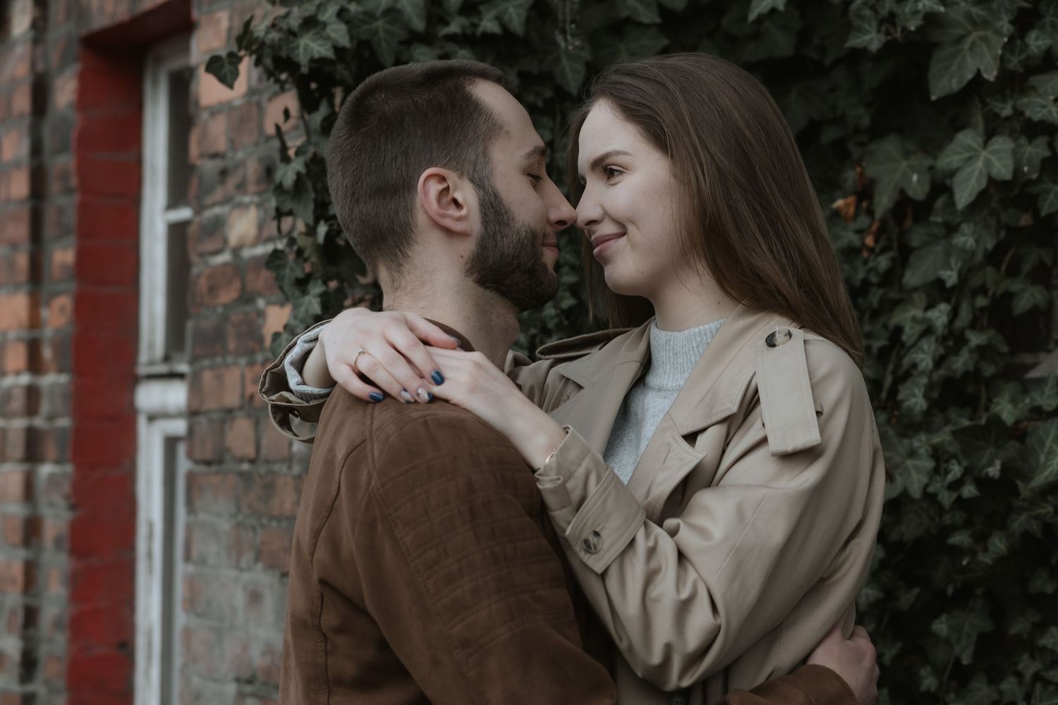 A romantic couple embraces against a brick wall with ivy, wearing brown and beige coats on an overcast day.