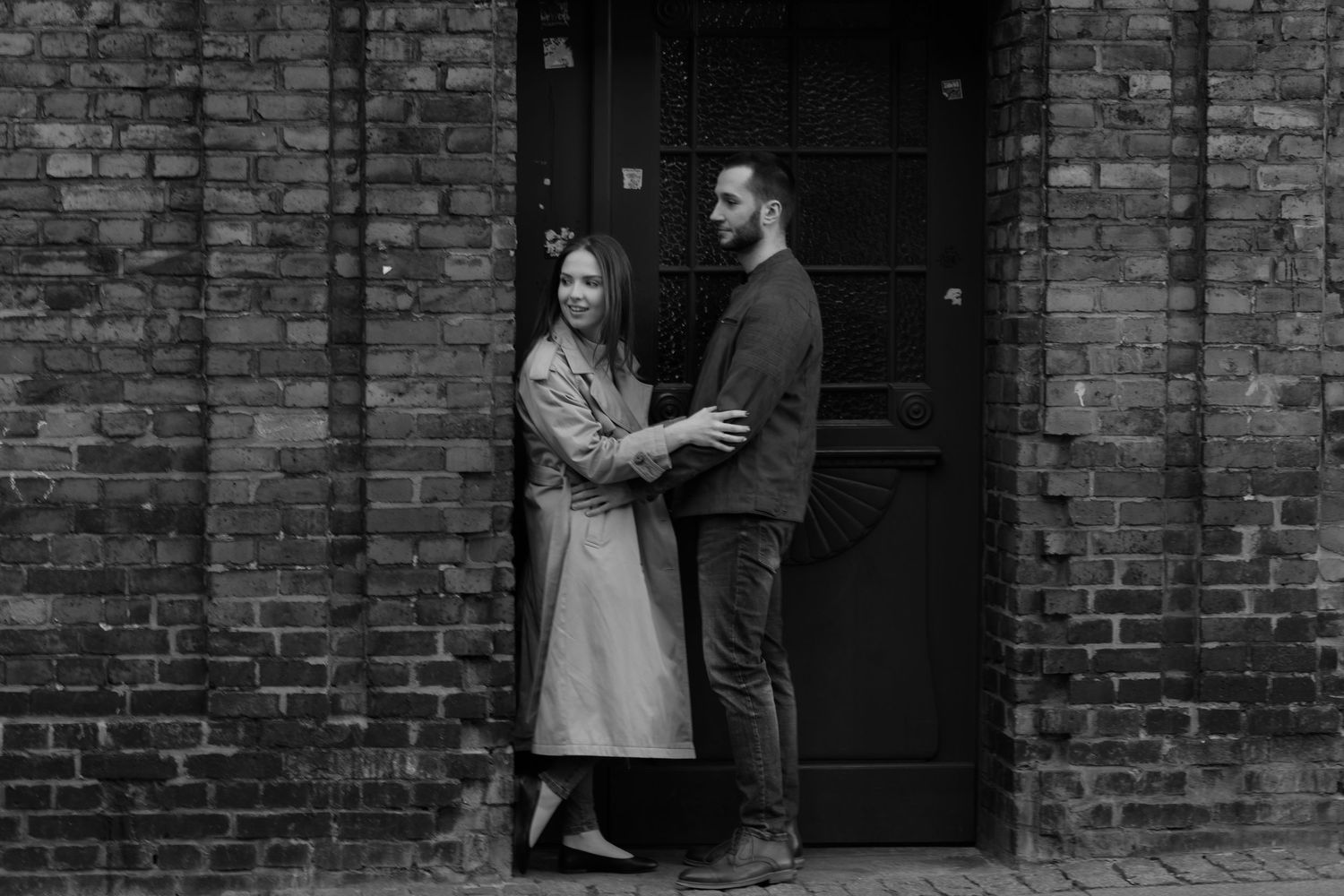 A couple shares an intimate moment while standing in front of a rustic brick wall and black door.