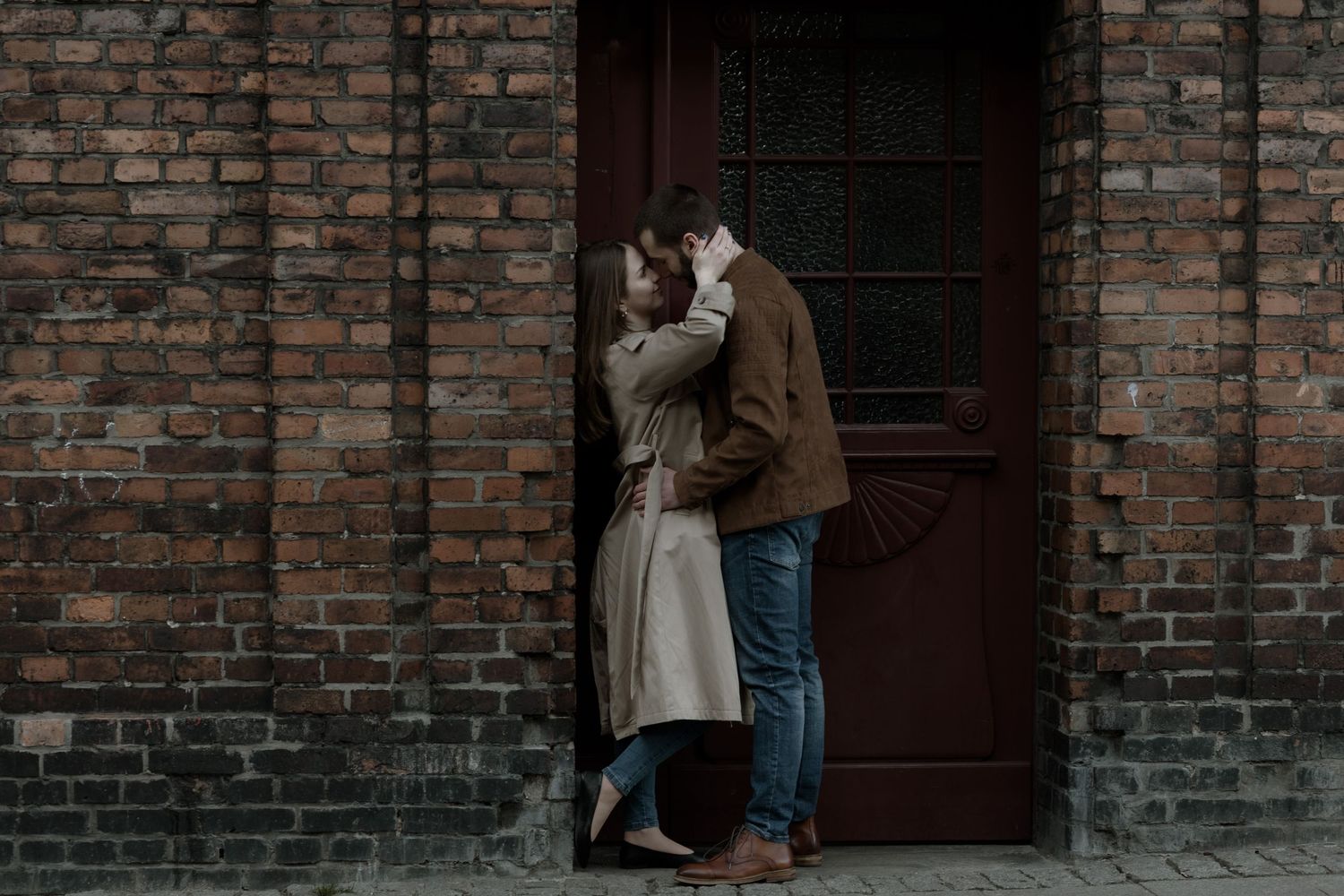 A romantic couple embraces in front of a rustic brick wall with a dark wooden door on a moody afternoon.