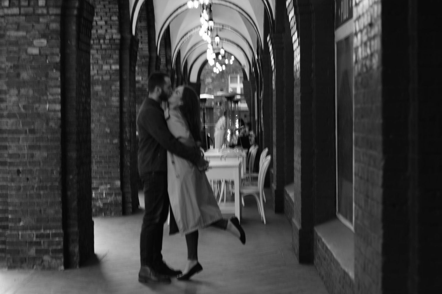 A couple stands close together in a long covered brick arcade corridor in this vintage black and white photograph.