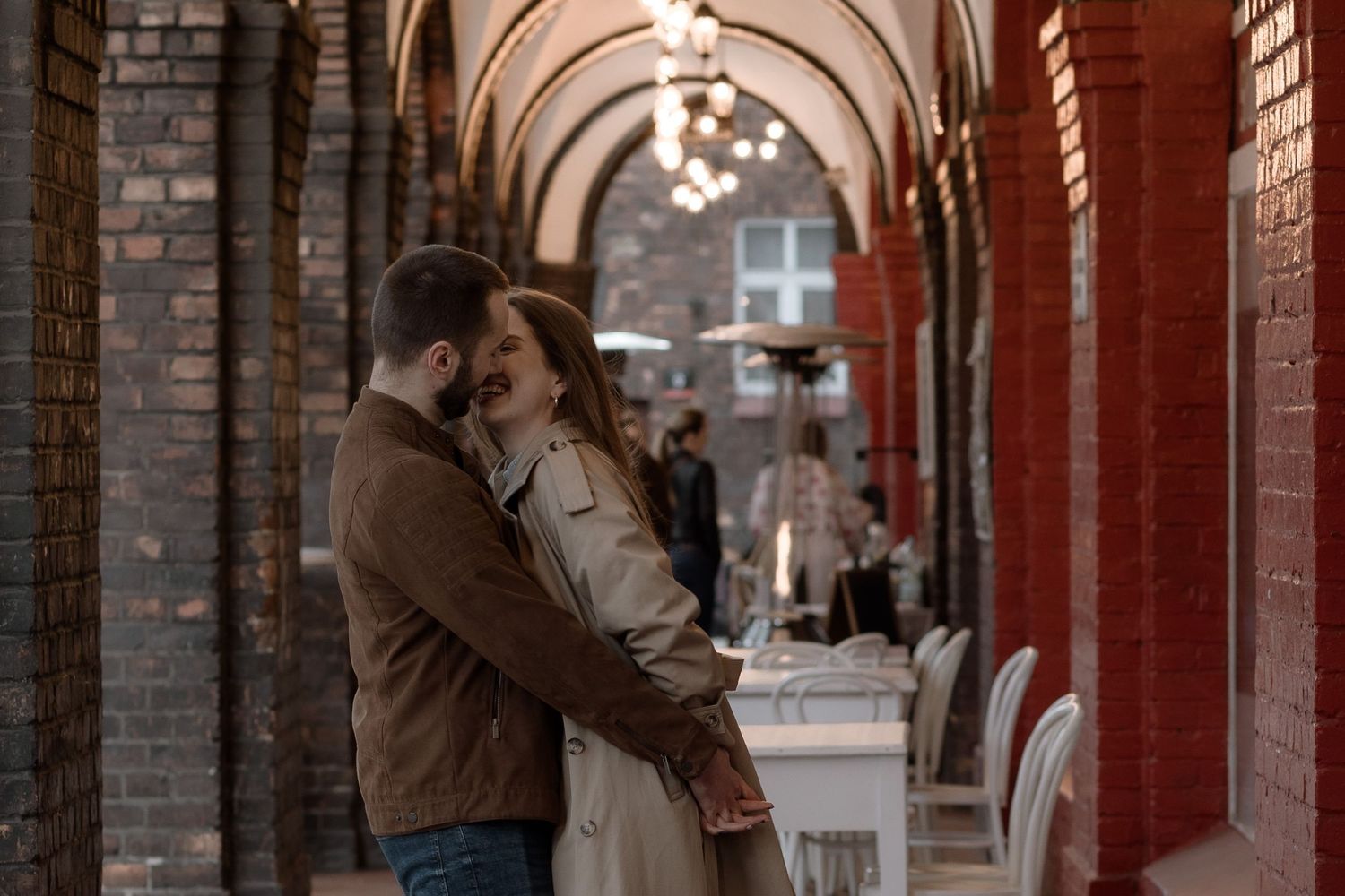 A couple embraces in a romantic arched brick corridor with decorative lighting and classical architecture.
