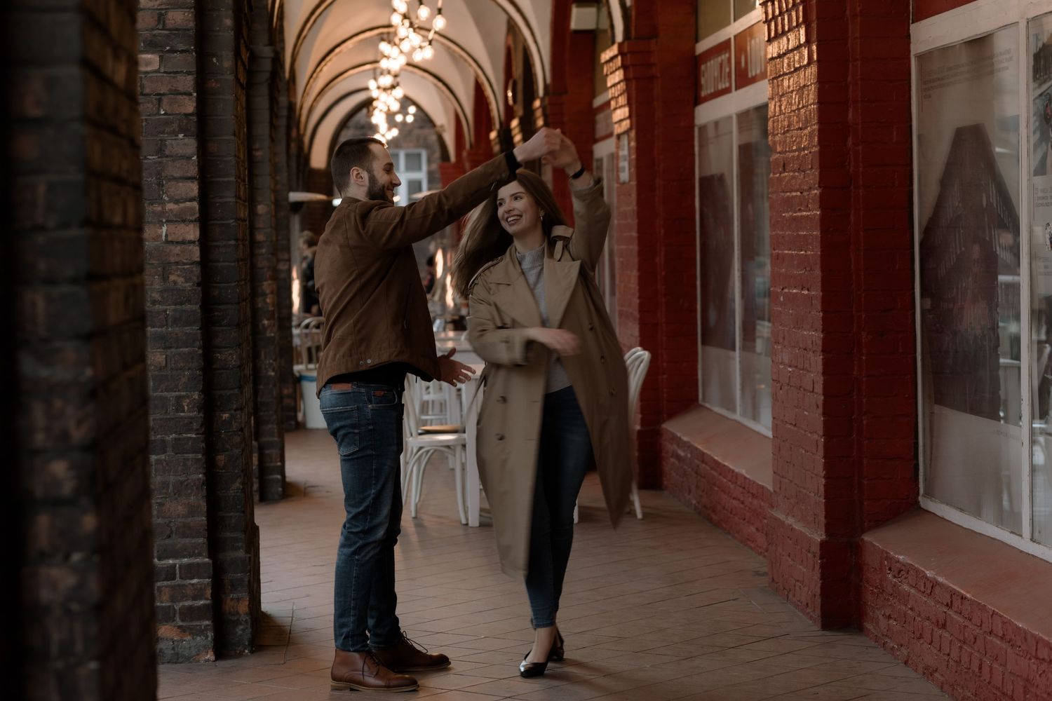 Two students chat while walking through an arched brick corridor with red walls and vintage window displays.