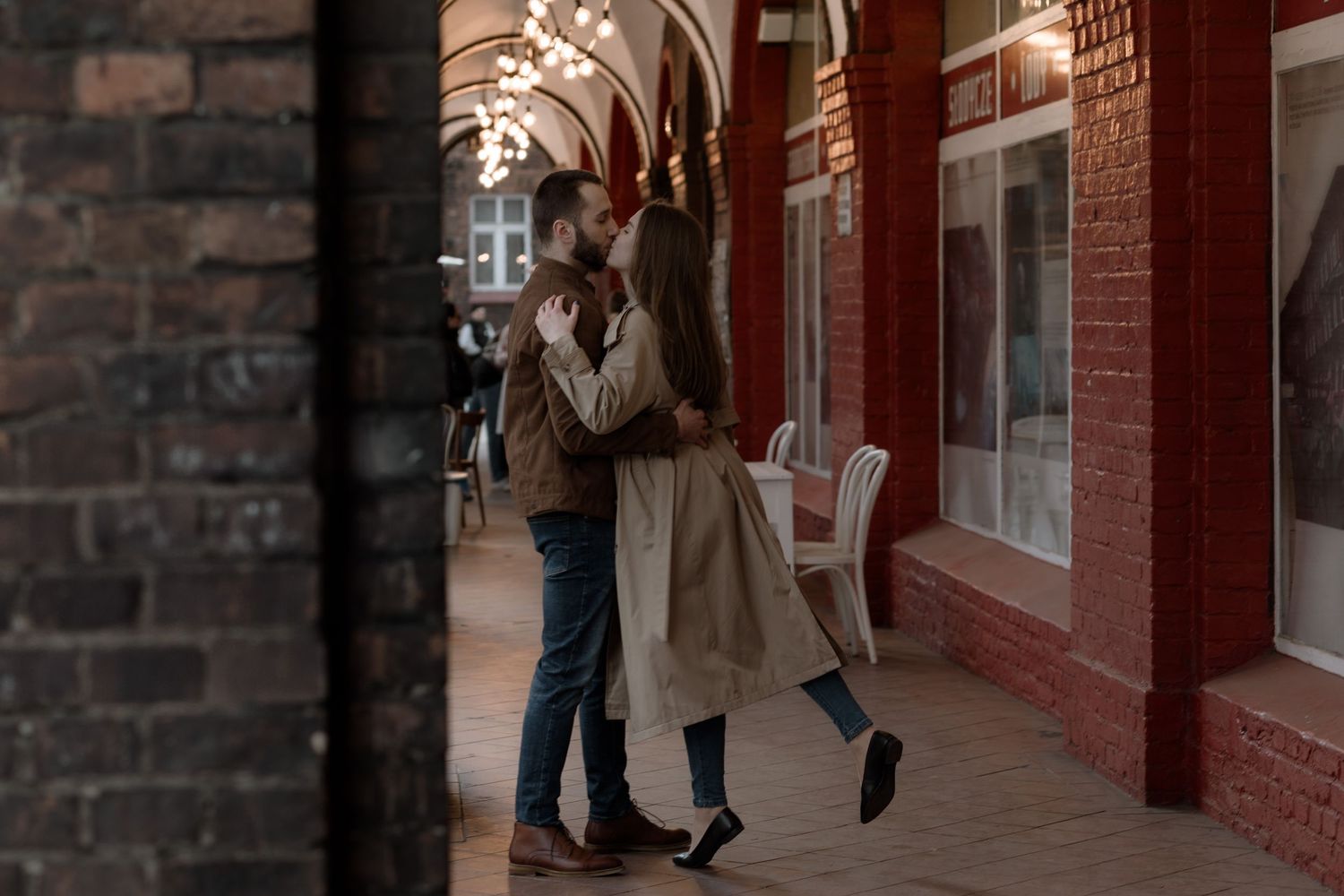 Two people share a romantic moment under an arched brick passageway during autumn weather.