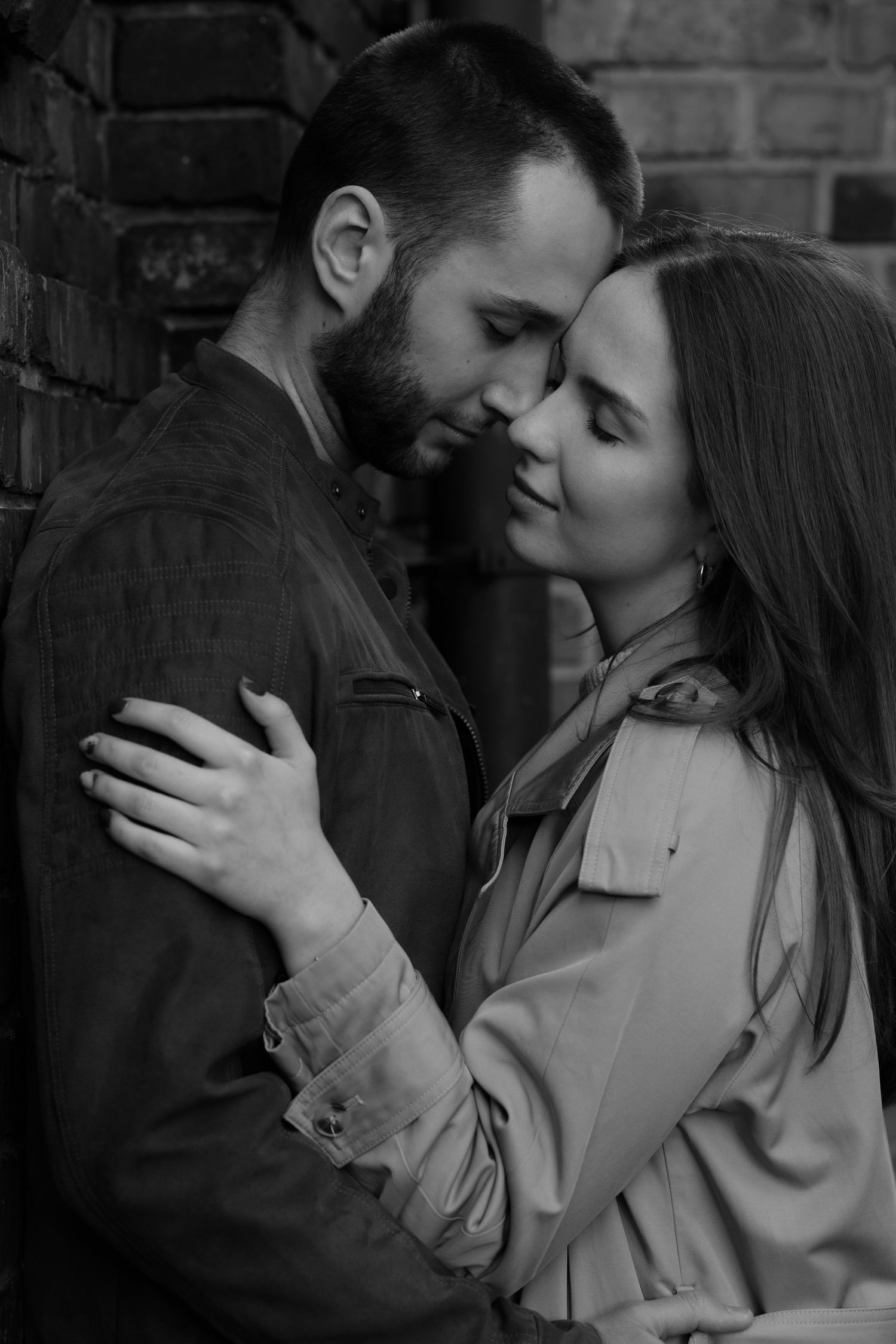 A romantic black and white portrait of a couple embracing against a brick wall.