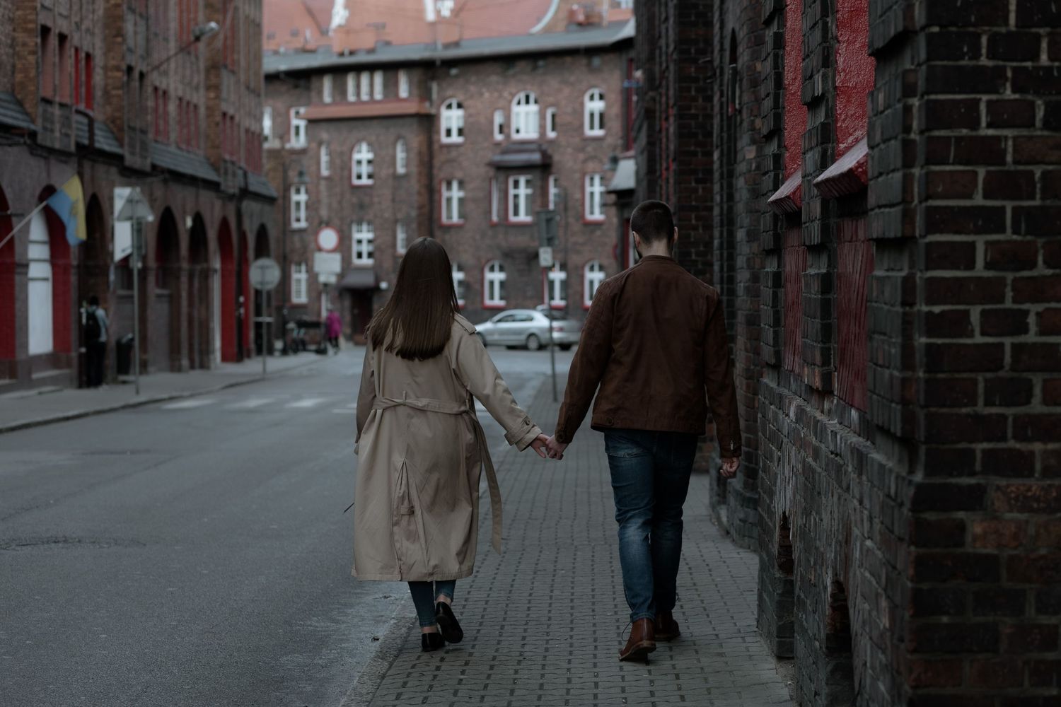 A couple walks hand in hand down a quiet cobblestone street with historic brick buildings in the background at dusk.