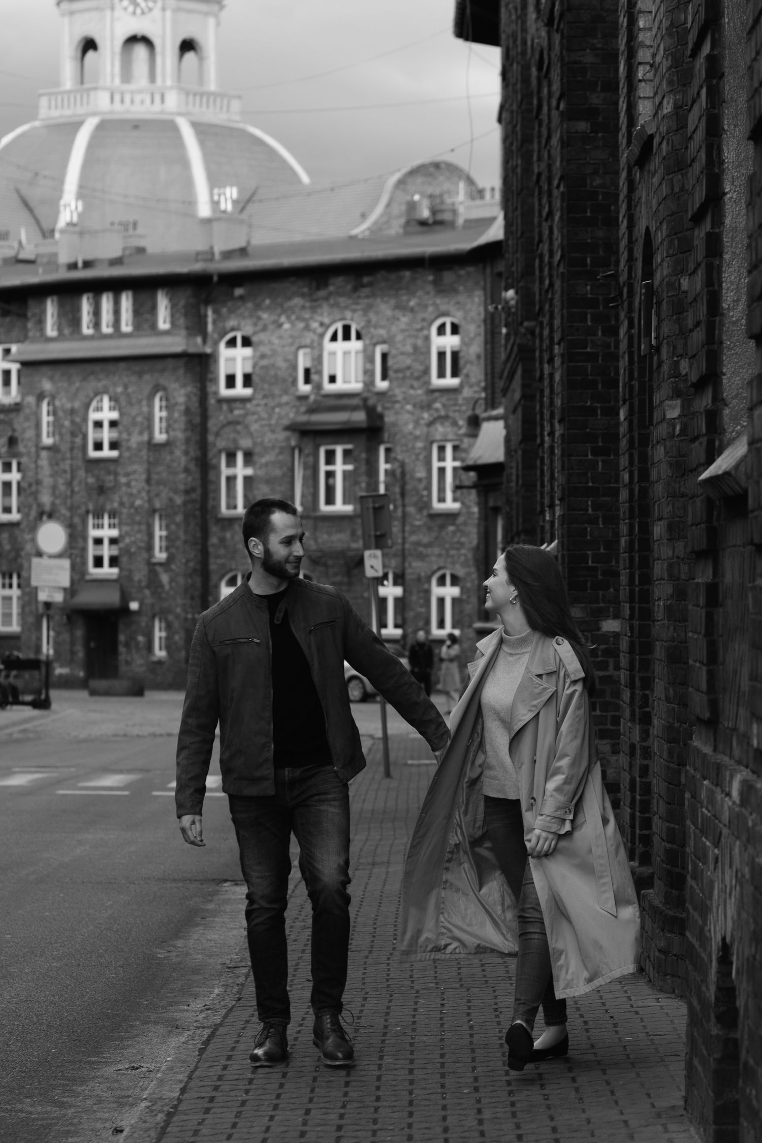 A couple walks together on a cobblestone street with historic architecture and a domed building in the background.