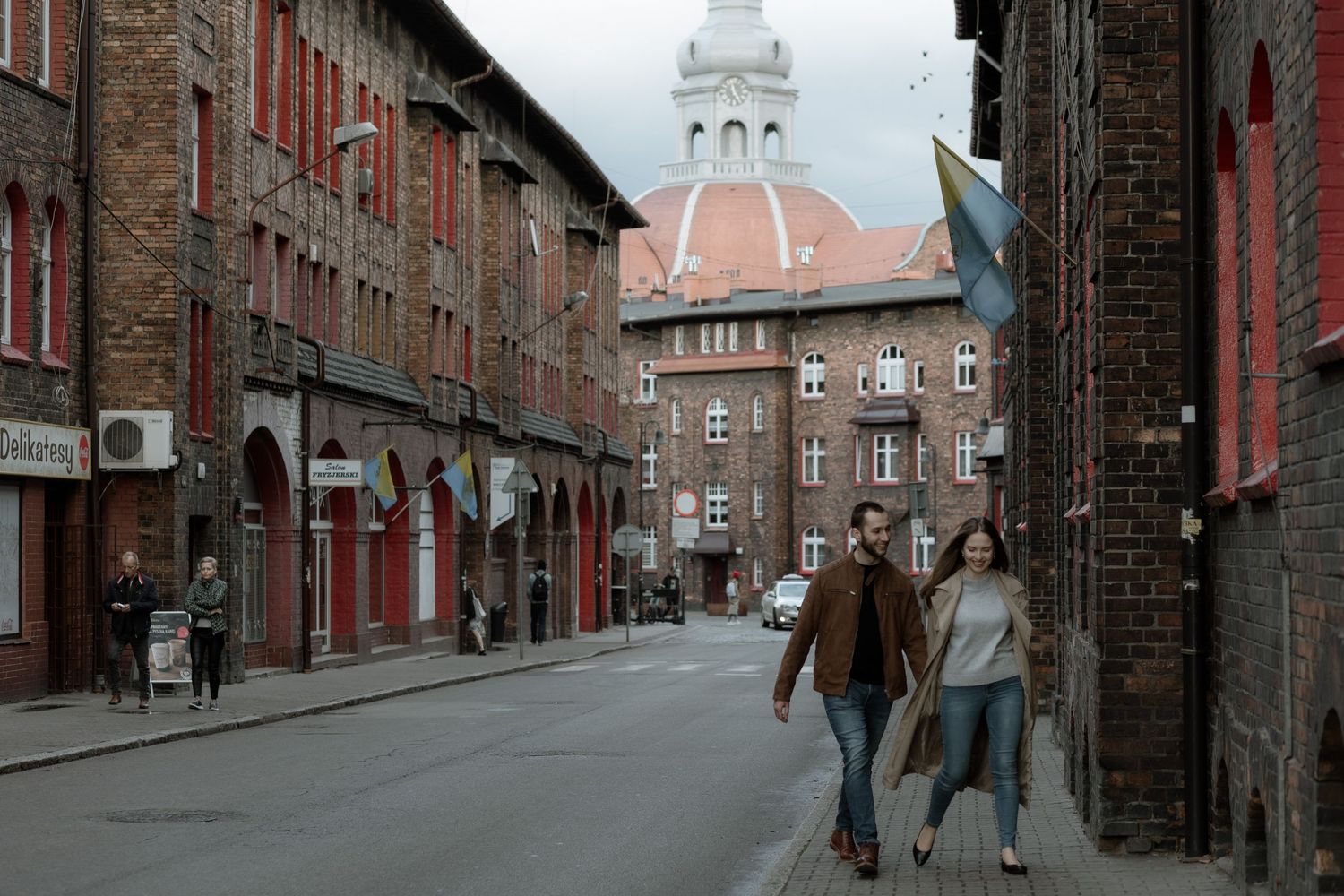 People walking down a historic European street with red brick buildings and a white clock tower in the distance.