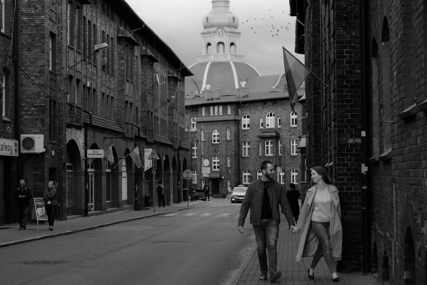 Vintage black and white street scene showing pedestrians walking past historic brick buildings with a decorative tower in distance.
