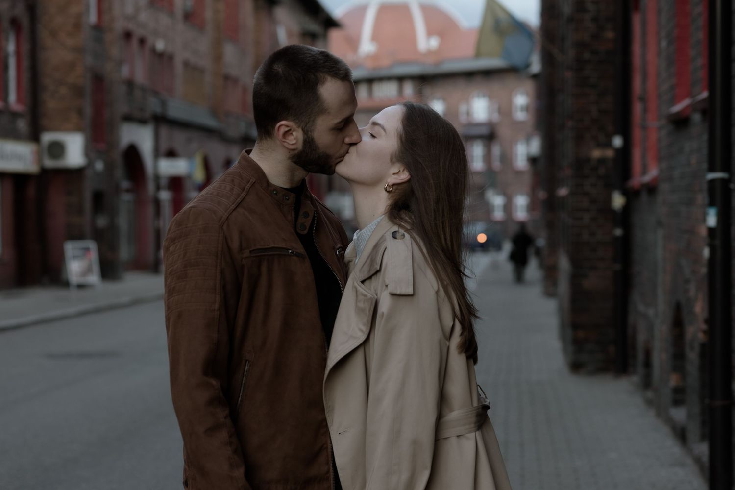 A couple shares a romantic kiss on a moody urban street with brick buildings and overcast skies in the background.
