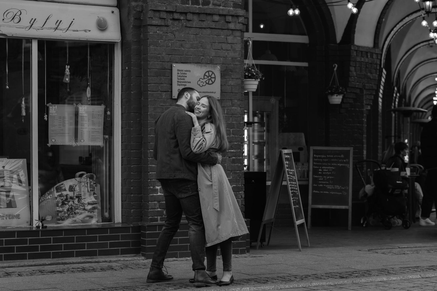 A couple embraces on a vintage city sidewalk outside a brick storefront in black and white.