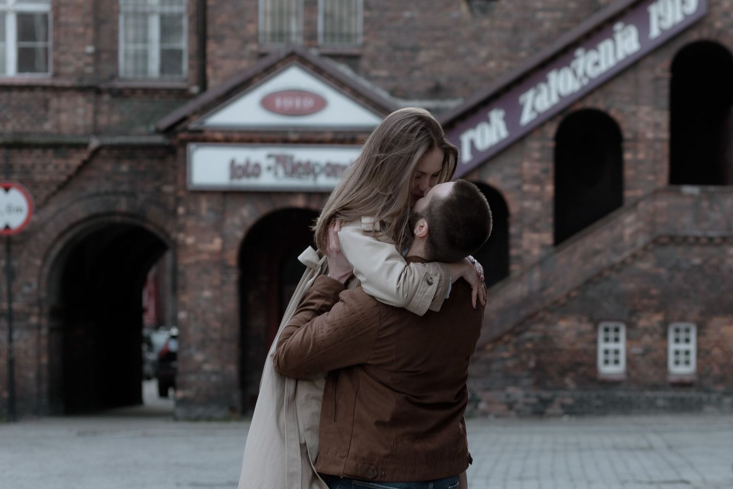 A couple embraces in a warm hug outside a historic brick building on a moody winter day.