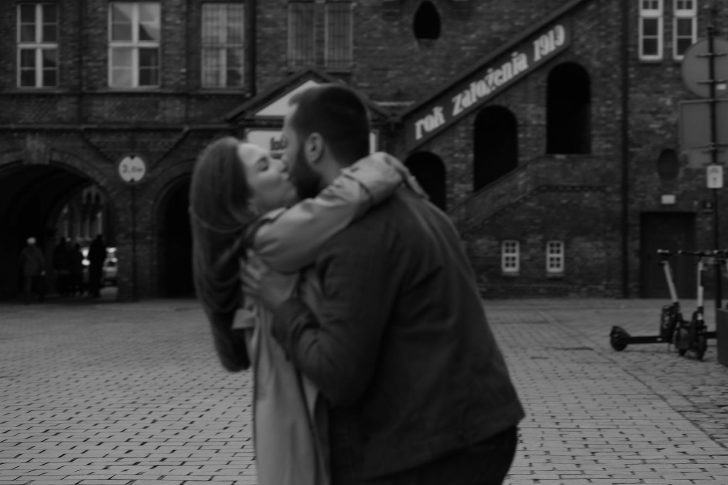 A tender romantic moment captured in black and white on a cobblestone courtyard with brick buildings in the background.