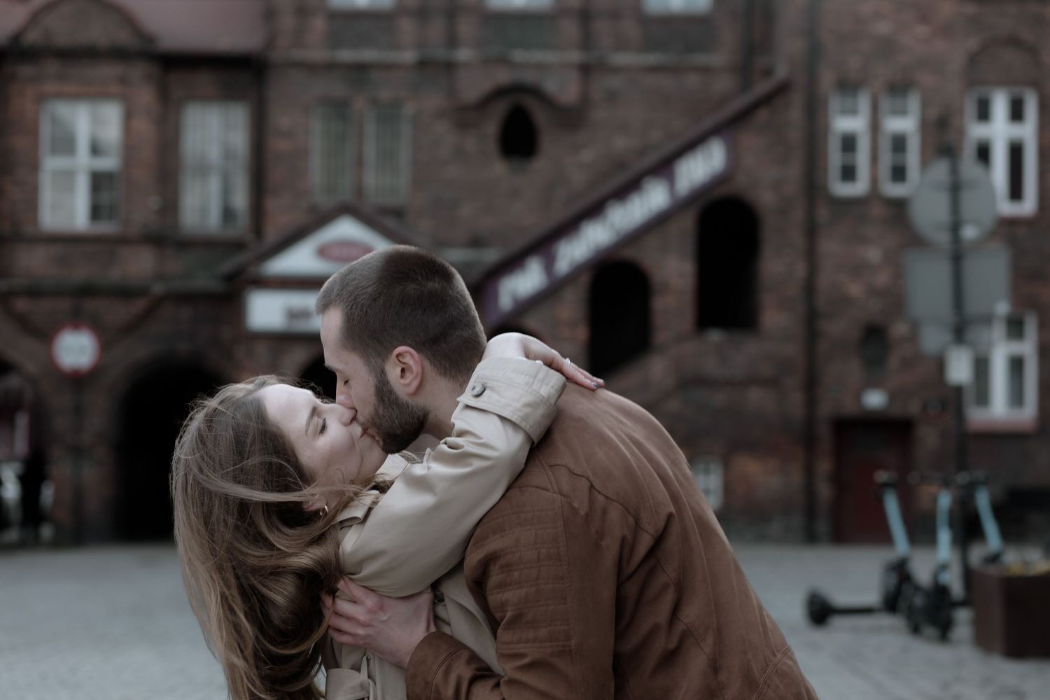A couple shares an intimate romantic kiss on a cobblestone street with old brick buildings in the background.
