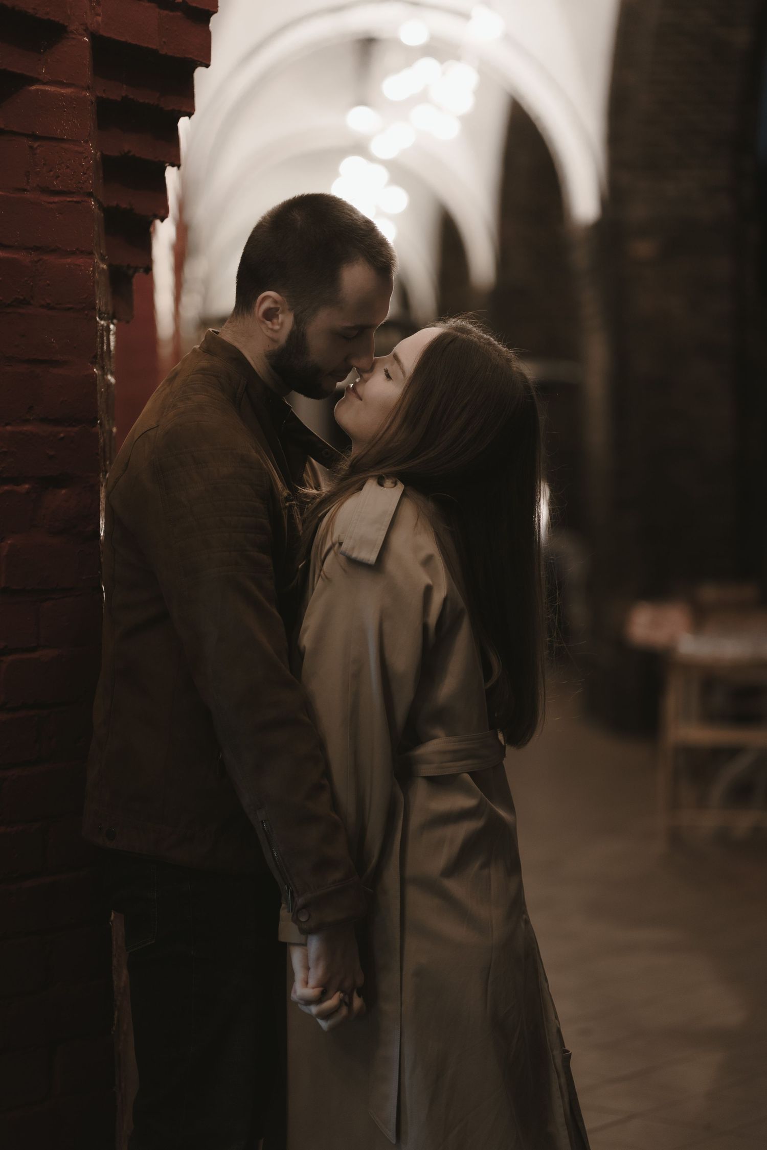 A romantic couple in trench coats share an intimate kiss against a brick wall under arched hallway lighting.