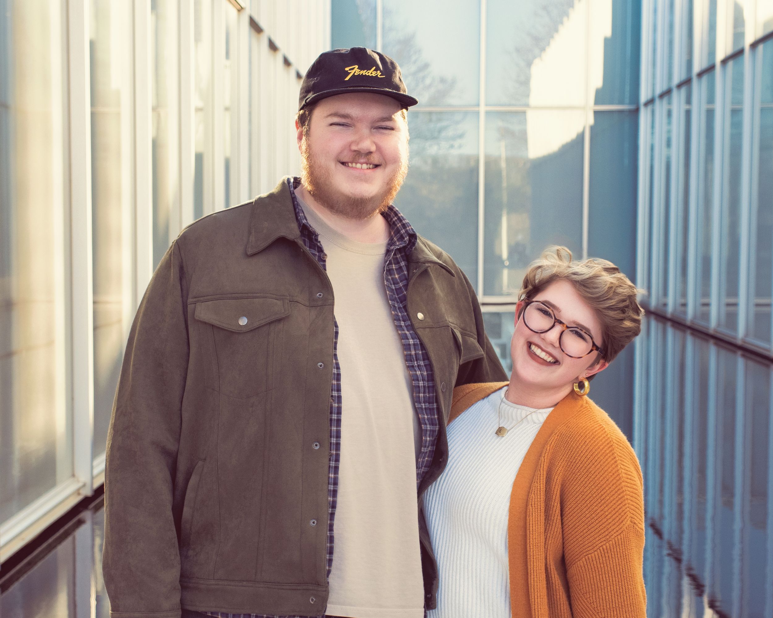 A couple smiles together while standing against a modern building's exterior during a sunny day.
