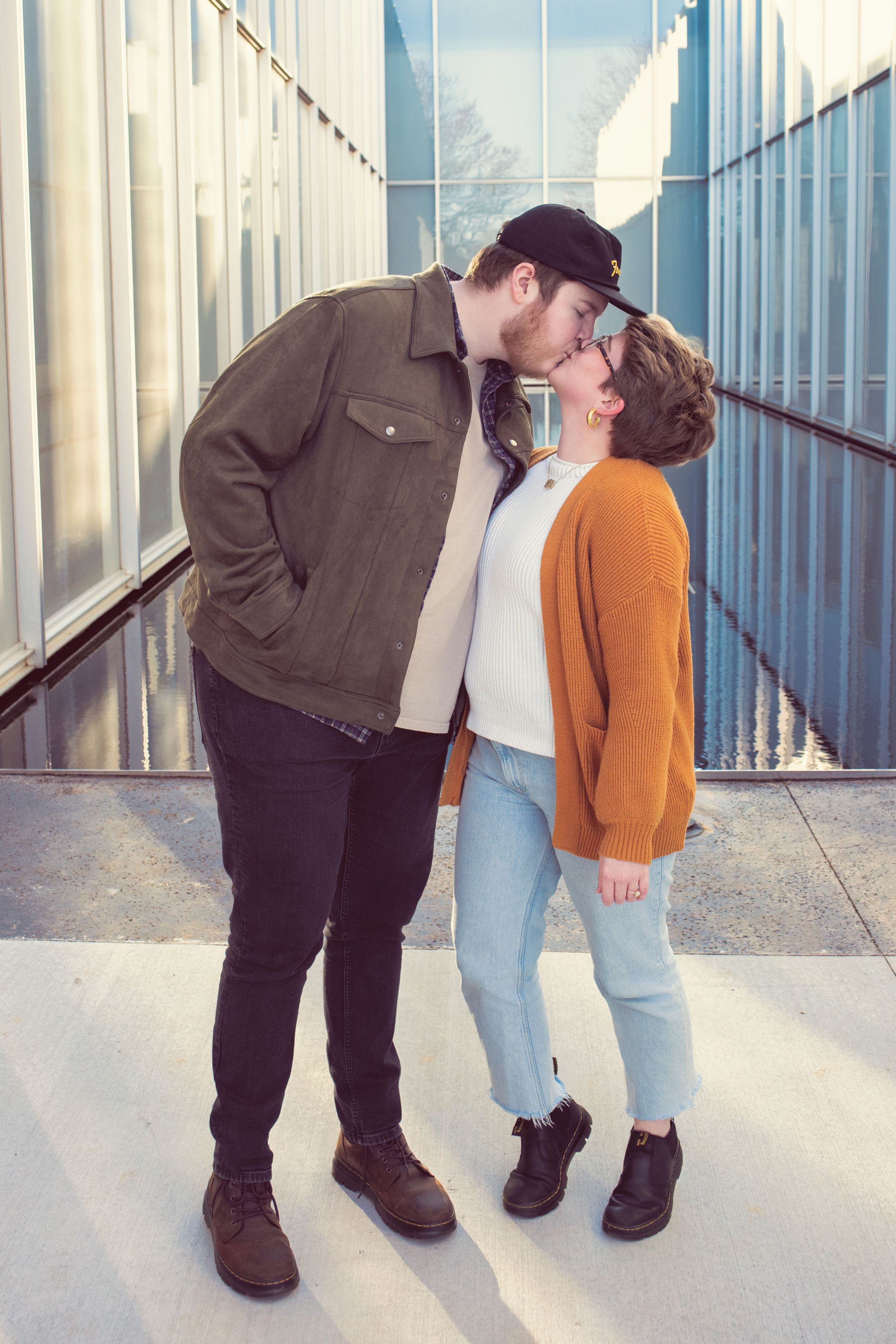 A couple in casual vintage-style clothing share a romantic kiss in front of a white industrial backdrop.