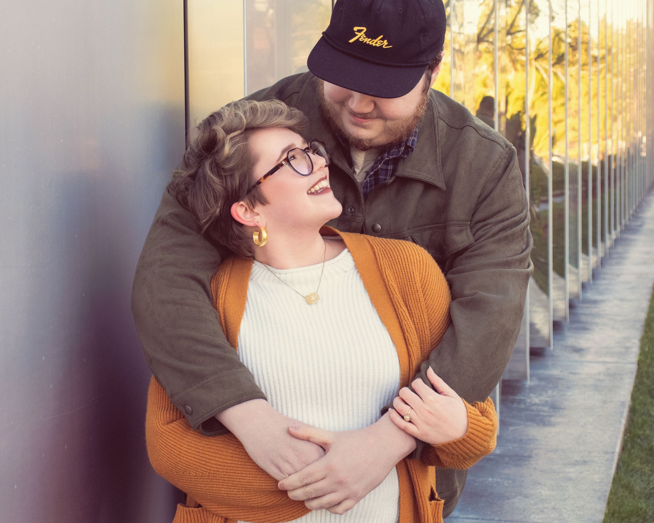 A couple embraces and laughs together while standing along a sunlit fence with shallow depth of field.