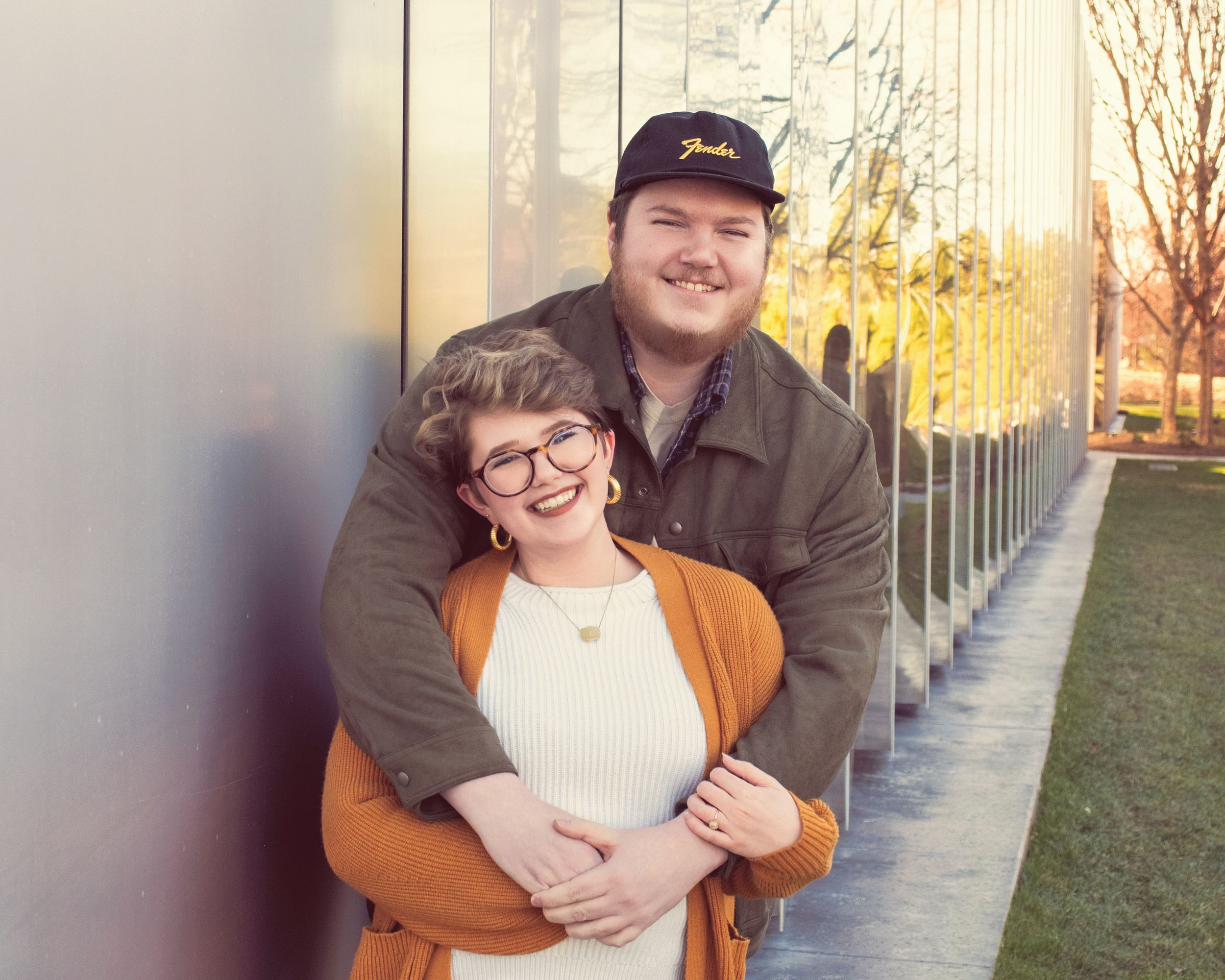 A couple embraces outdoors on a path lined with metal posts during golden hour sunlight.