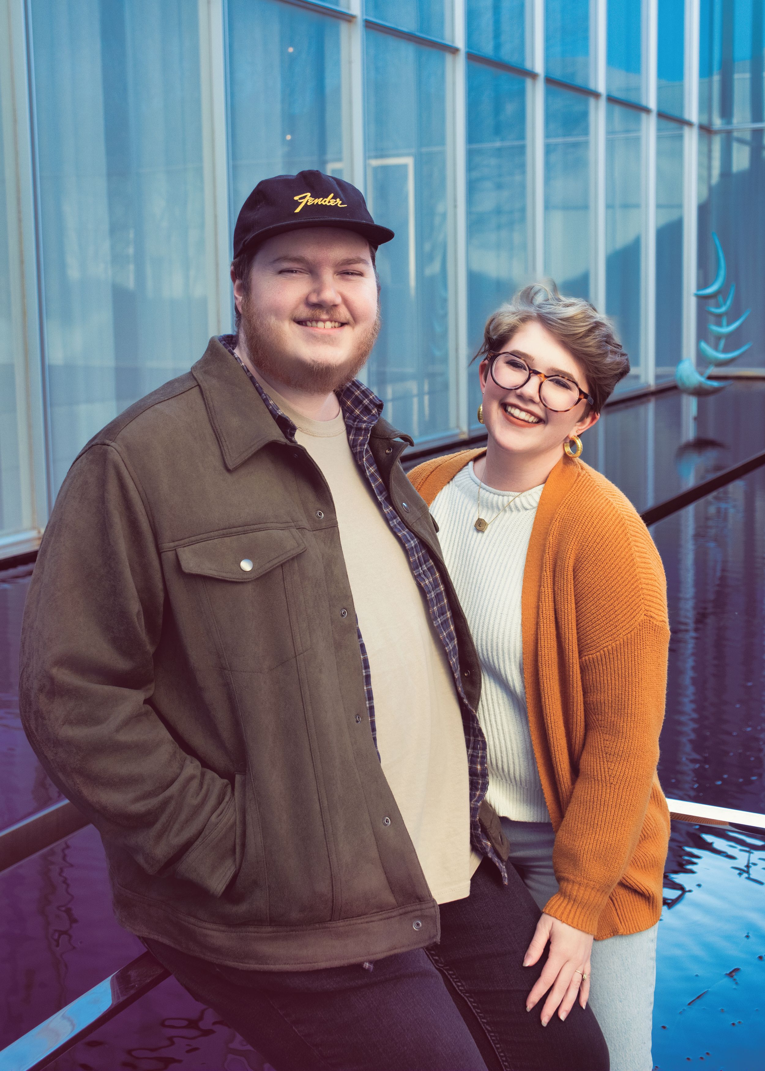 A couple stands smiling together against a blue industrial wall on a bright day.