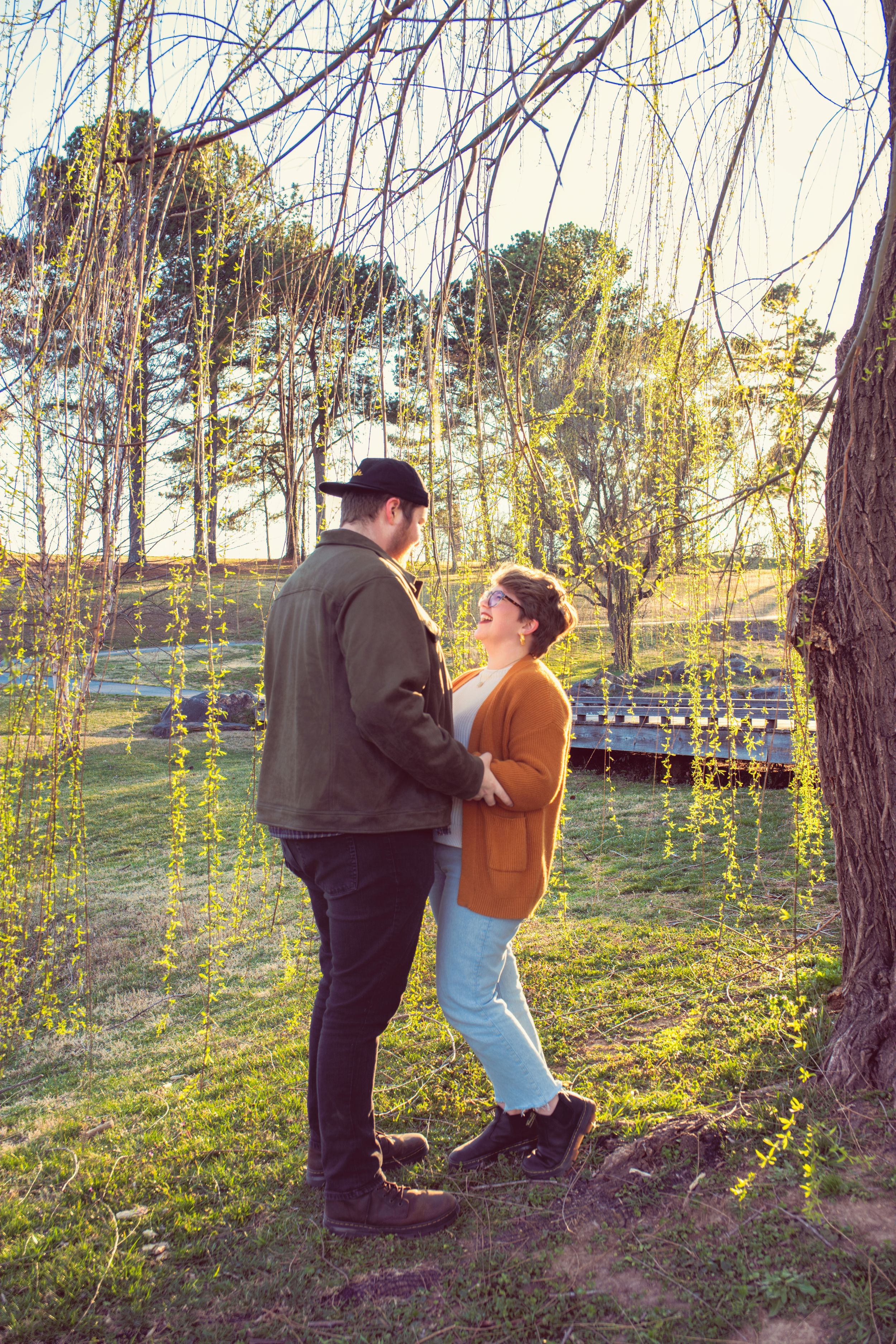 Two figures share an intimate moment under drooping willow branches in golden afternoon light.
