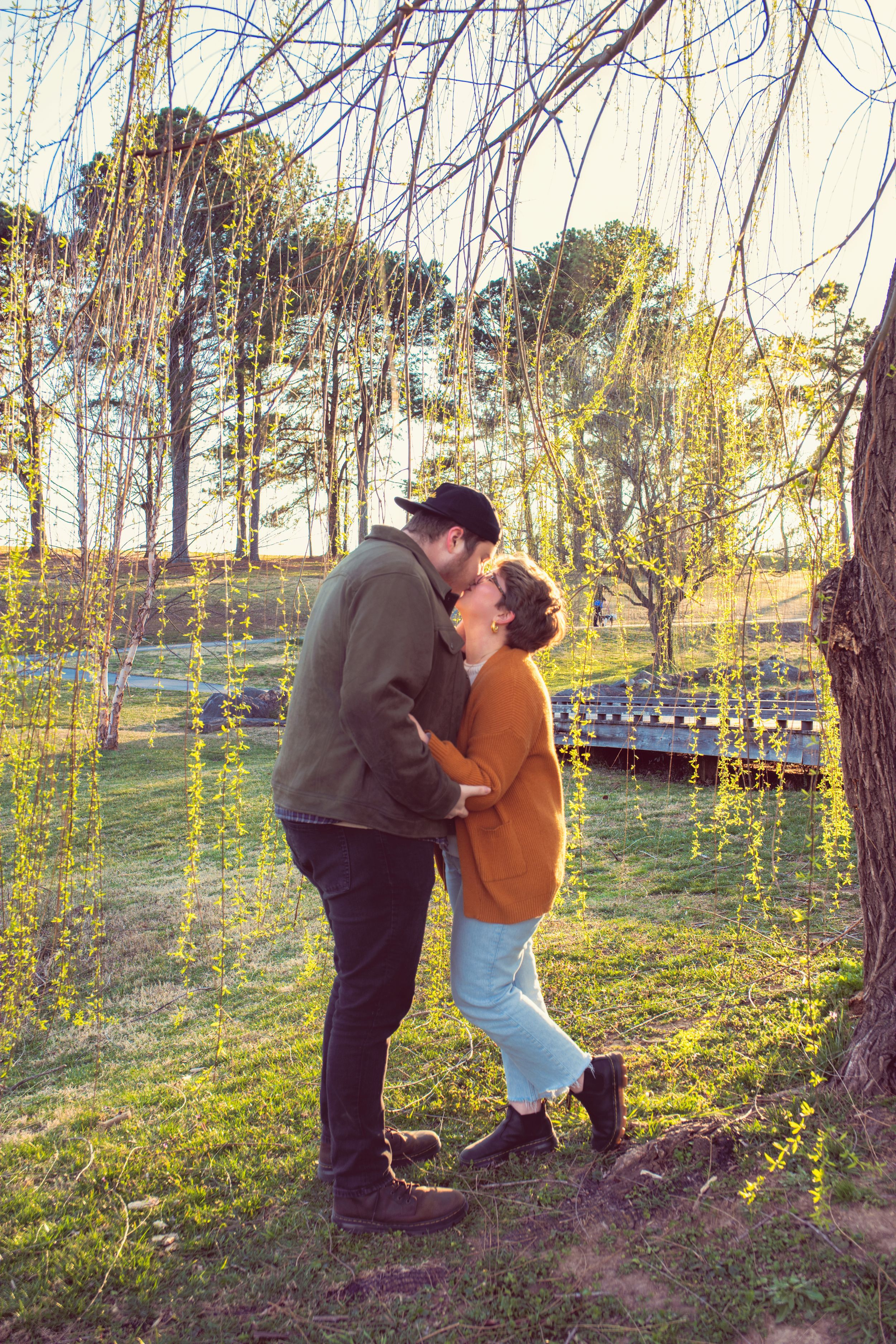 A couple shares a romantic moment under a weeping willow tree on a sunny afternoon in a scenic park setting.