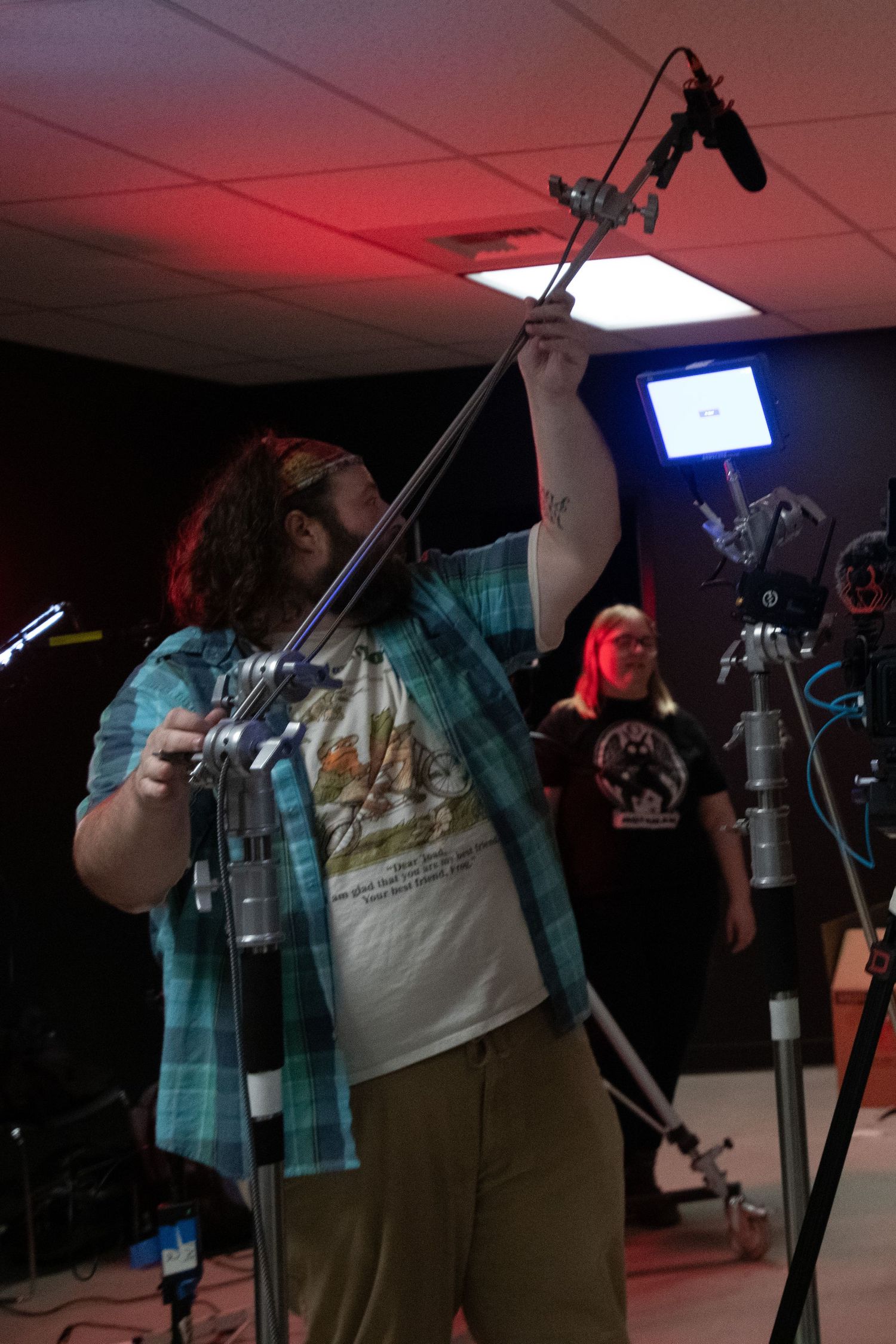 Man holding a boom microphone pole on a film set with red lighting and camera equipment in background.
