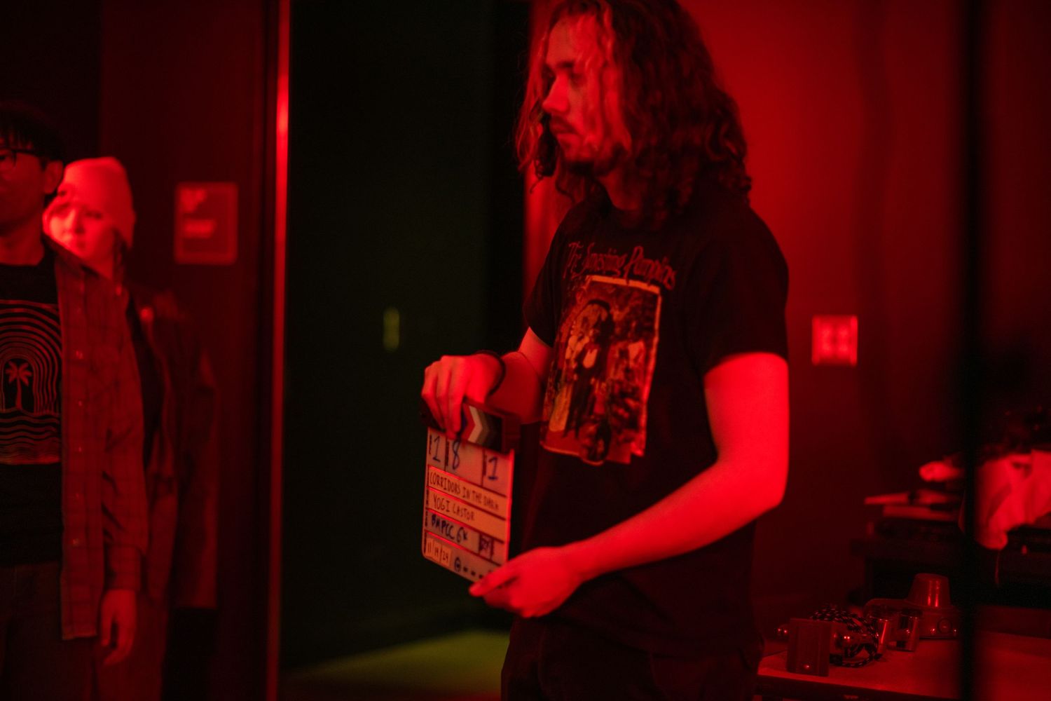 Long-haired man holding a film clapperboard on a red-lit movie set.