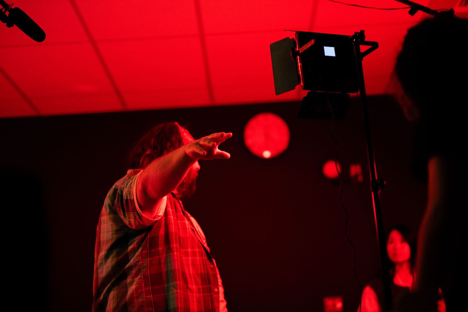 Person in plaid shirt gesturing on a red-lit film set with professional lighting equipment.