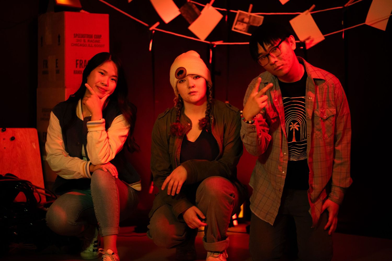 Three people posing together under red lighting on a film set decorated with hanging polaroid photos.